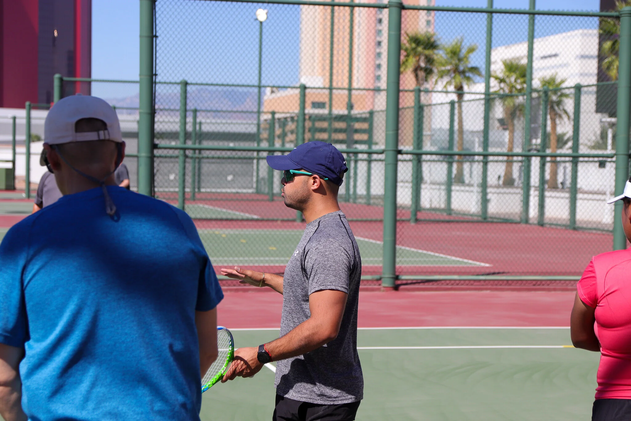 A man in a gray shirt, blue cap, and sunglasses appears to be instructing or coaching on an outdoor tennis court, with other players partially visible nearby. The court is surrounded by a tall green fence, with a cityscape of tall buildings and palm