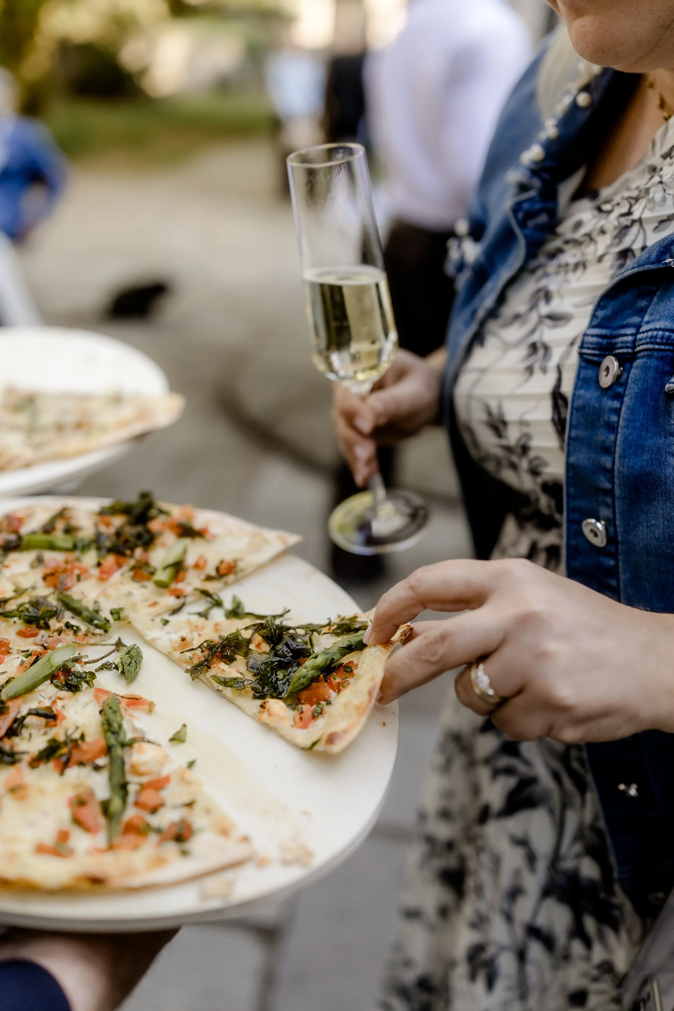 Person beim Servieren von Pizzastücken mit Tomaten, Spargel und Grünzeug auf einem Pizza-Tablett, während eine andere Person ein Glas Sekt hält.