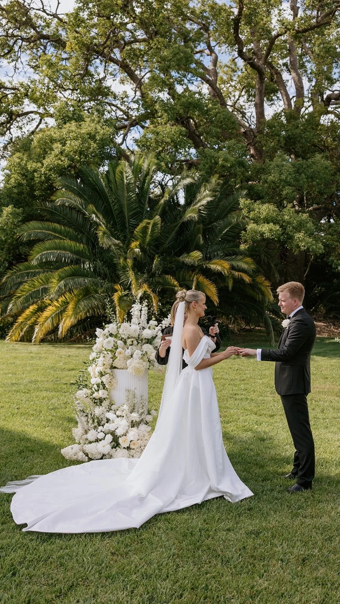 Bride and groom exchange rings at an outdoor wedding ceremony on a lawn with a large palm tree and lush greenery in the background.