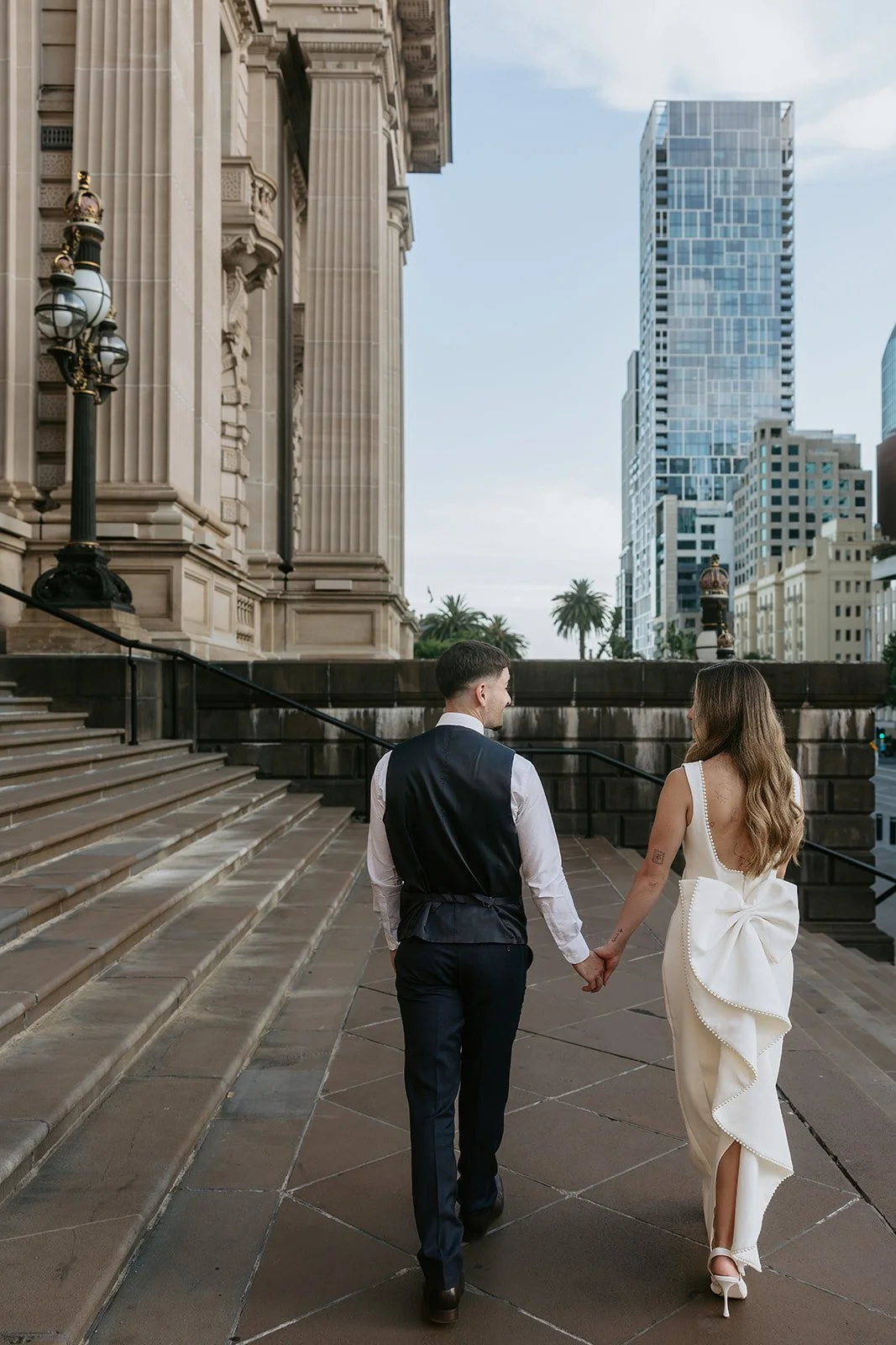 A couple dressed in formal wedding attire holding hands and walking up the steps outside a historic building with ornate architecture, large columns, and city skyscrapers in the background.