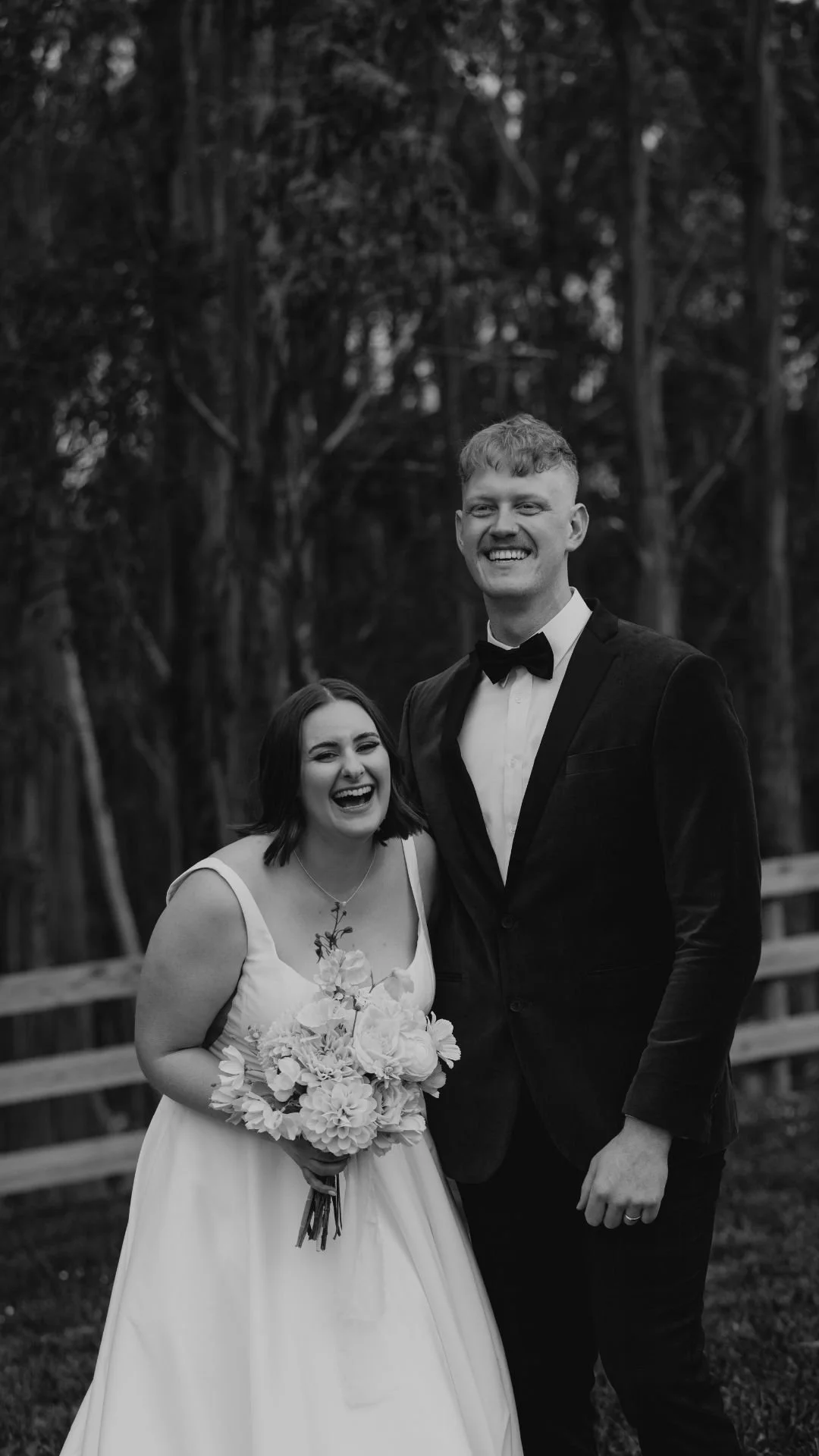 A black-and-white photo of a smiling bride holding a bouquet of flowers and a groom dressed in a tuxedo with a bow tie, standing outdoors with trees in the background.