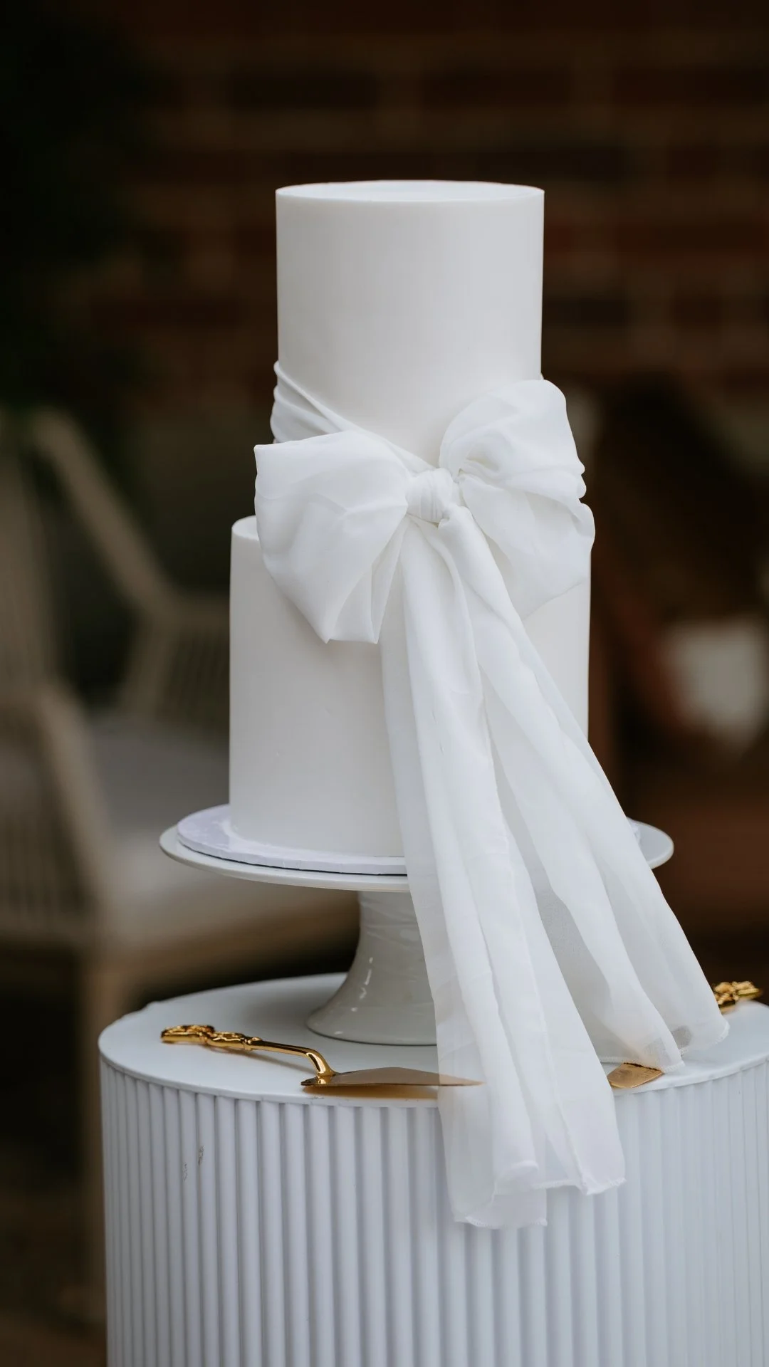 Two-tier white wedding cake with a white ribbon bow decoration, placed on a white cake stand with a gold cake knife beside it.