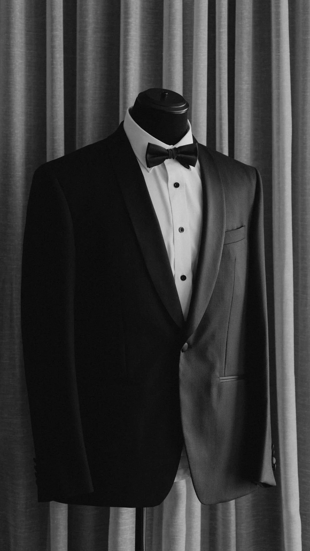 A mannequin dressed in a black tuxedo with a white shirt and black bow tie, standing in front of a curtain.