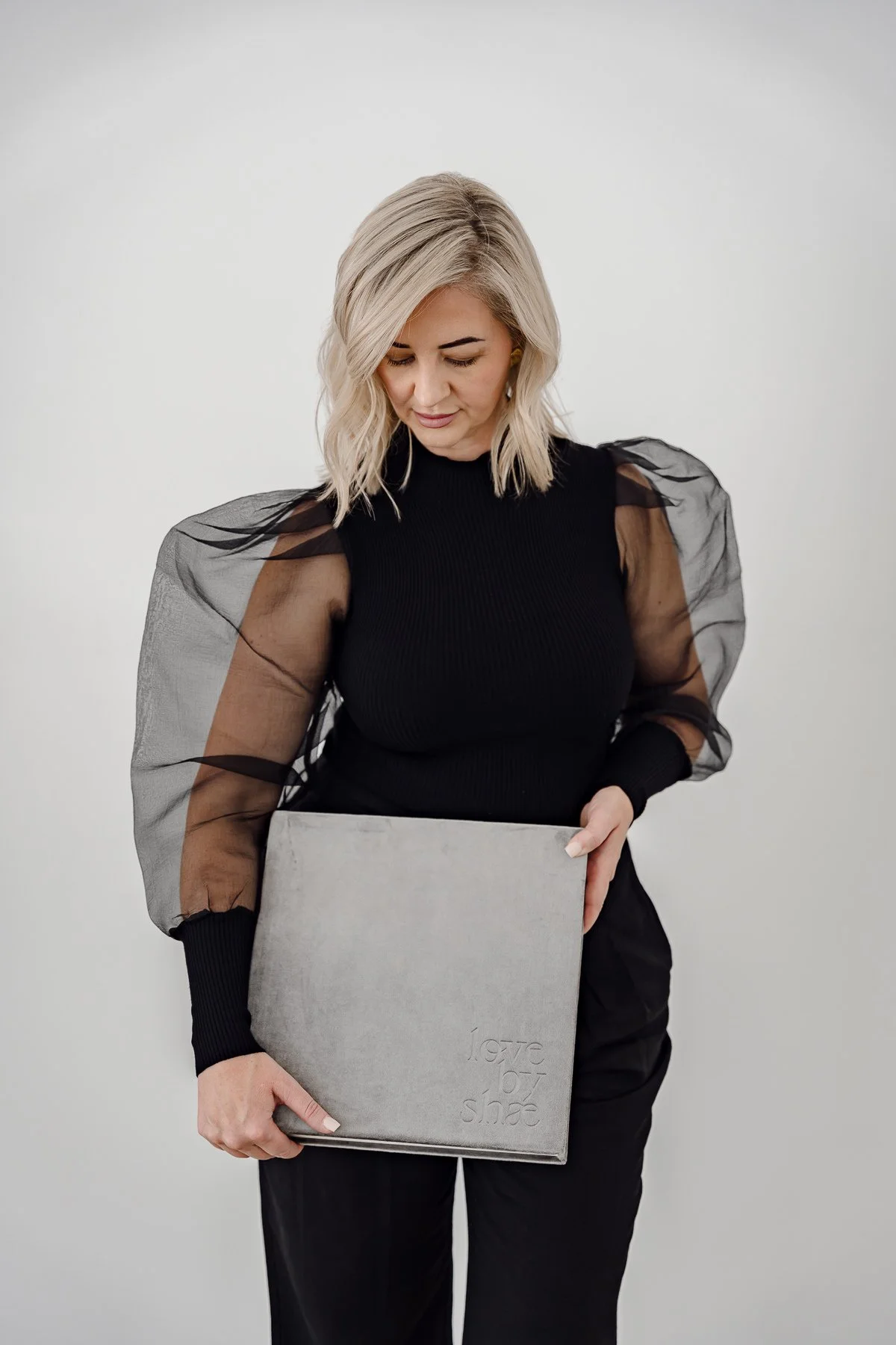 A woman with blonde hair wearing a black top with sheer, puffy sleeves, holding a gray book or folder.