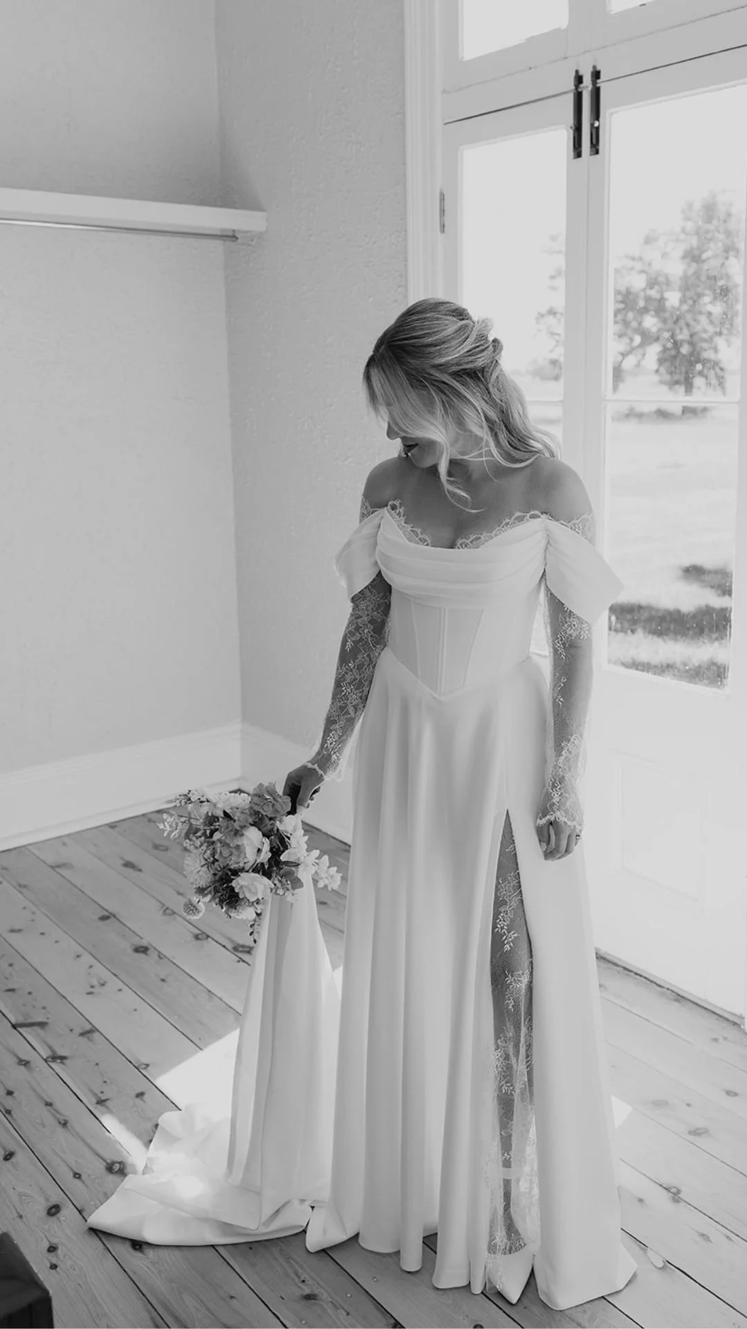 A woman in a bridal gown holding a bouquet, standing in a room with wooden floors, near a large window, in black and white.