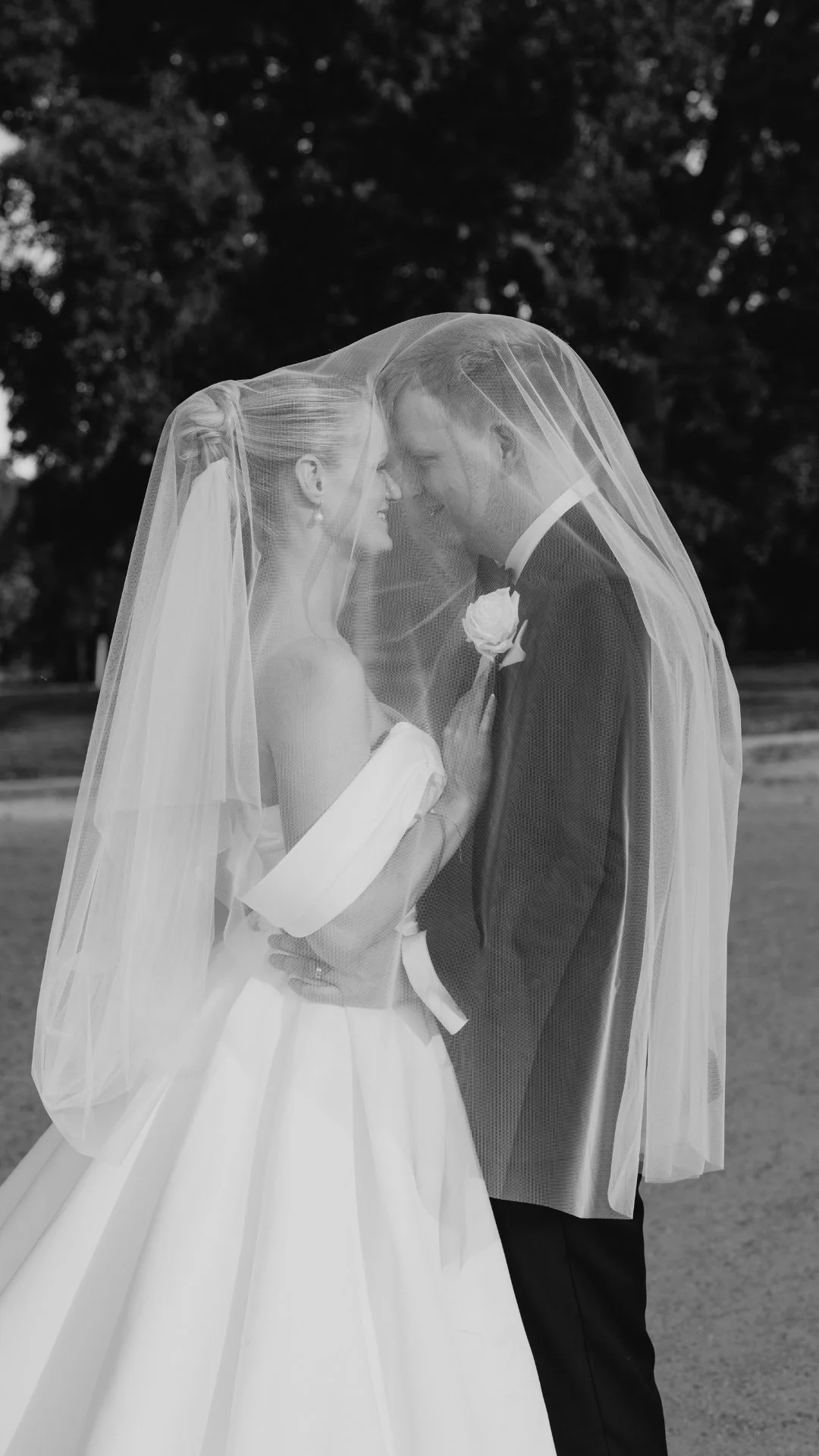 A bride and groom under a wedding veil, touching foreheads and smiling outdoors.