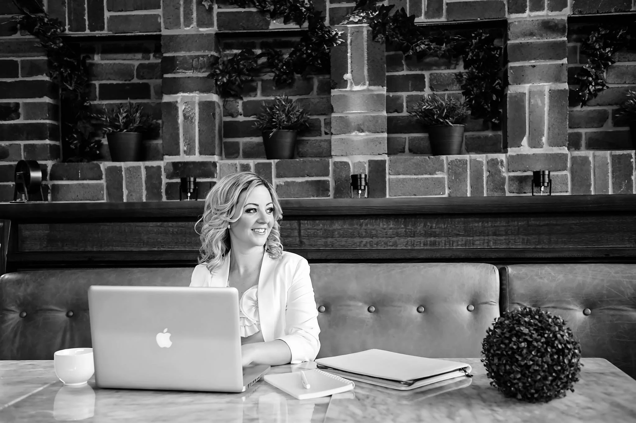 A woman with blonde hair and a white blouse sitting at a wooden table using a laptop in a modern office with an exposed brick wall and plants in the background.