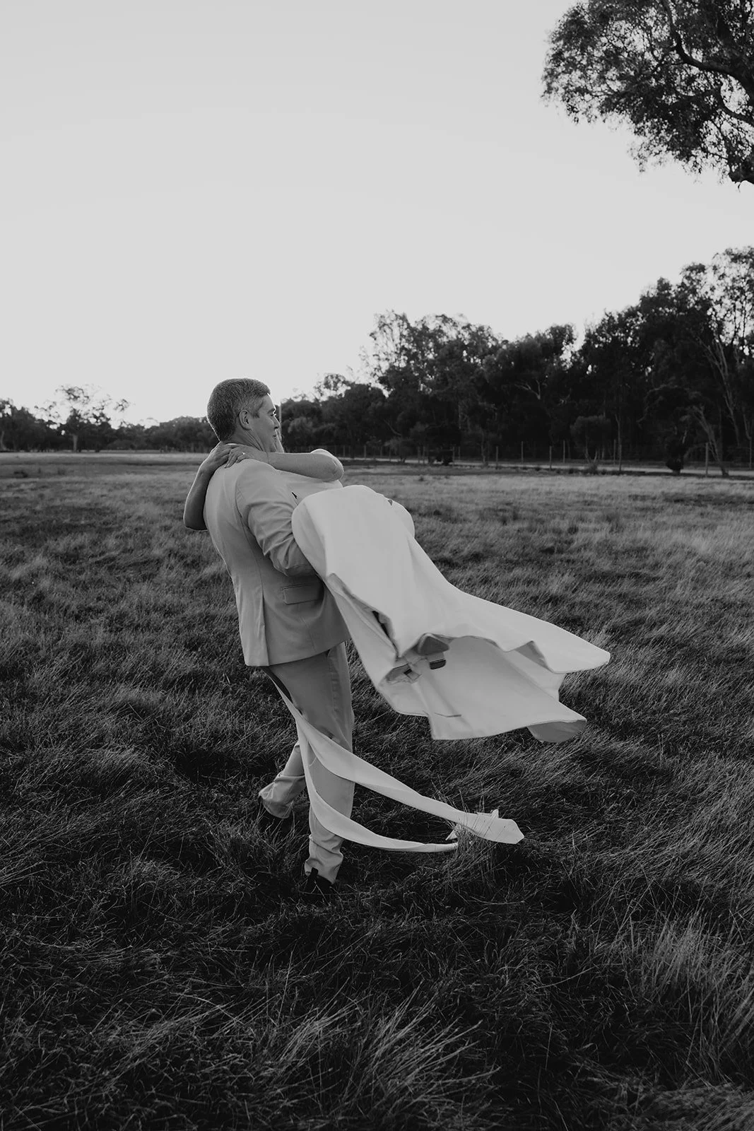 A black and white photo of a man in a light suit carrying a woman in a wedding dress across an open grassy field, with trees in the background.
