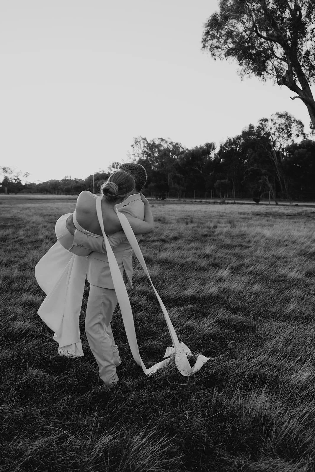 A black and white photo of a couple in wedding attire embracing in a grassy field with trees in the distance.