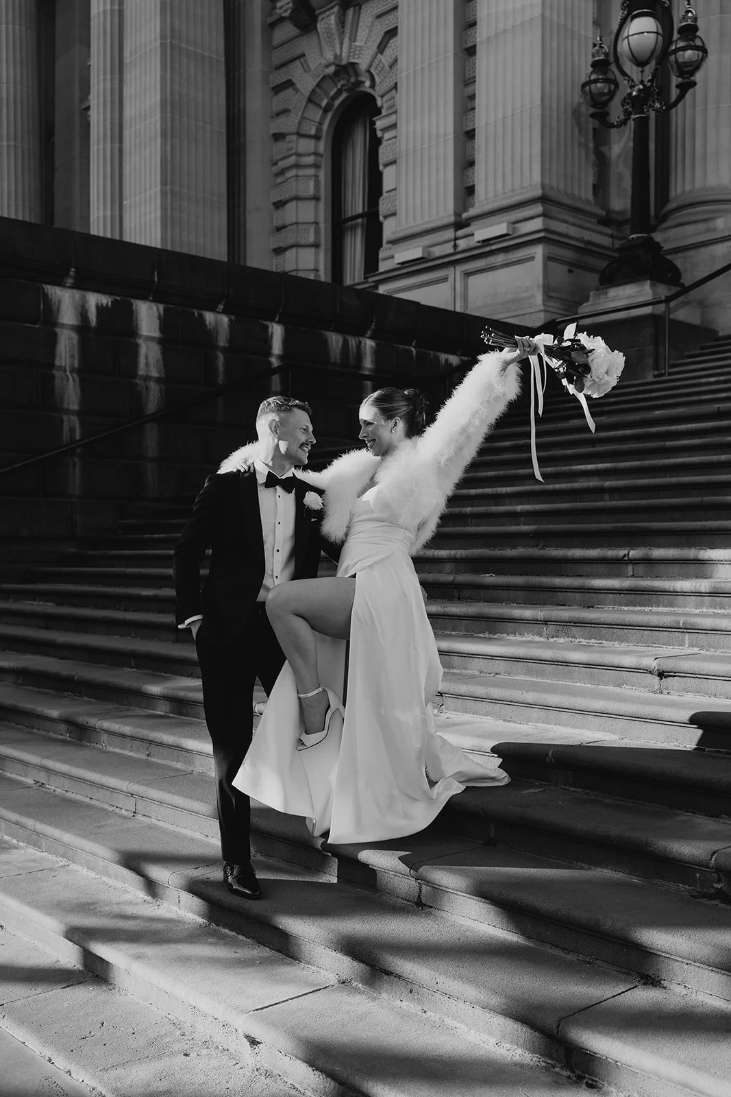 Black and white photo of a wedding couple on steps outside a historic building. The bride is holding a bouquet, raising her arm, and smiling at the groom. The groom is dressed in a tuxedo, smiling back at her.