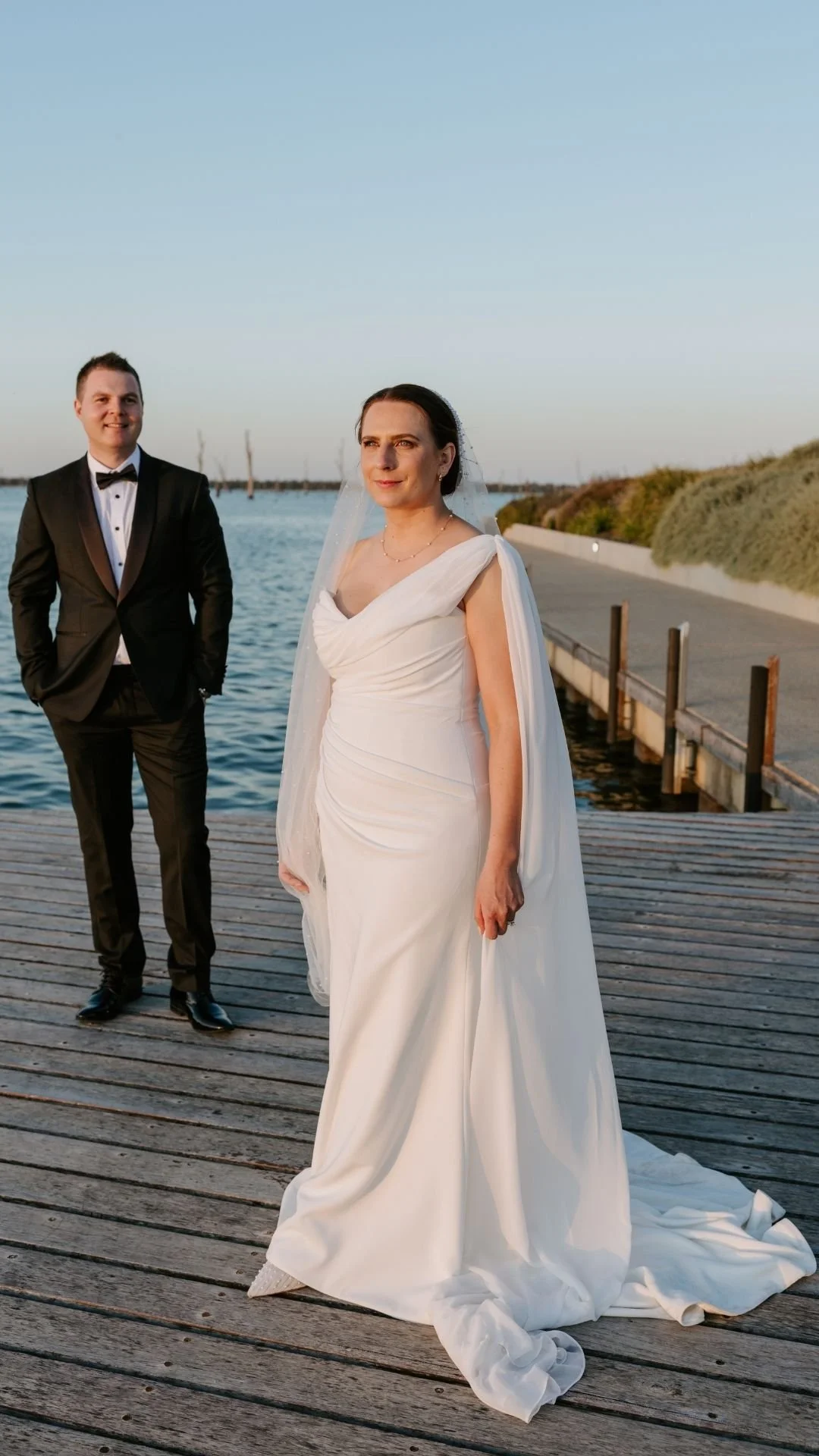 A bride in a white wedding gown and a groom in a black tuxedo standing on a wooden dock by the water during sunset.