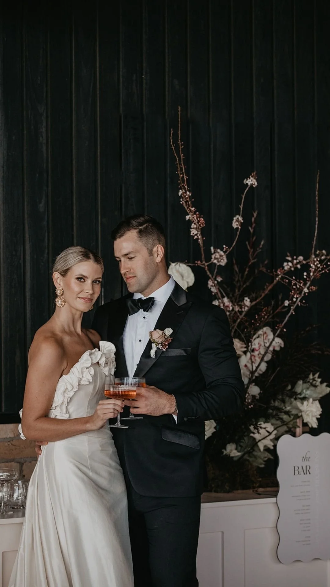 A bride and groom are celebrating at their wedding, holding glasses of champagne in front of a dark wall with floral arrangements.