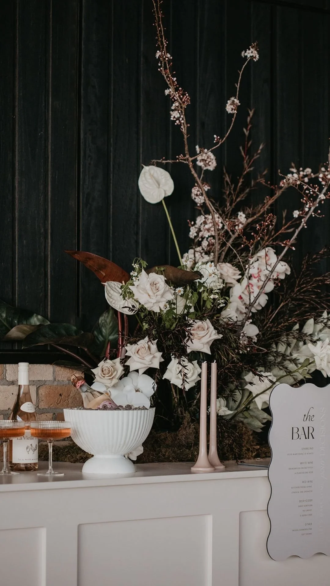 A floral arrangement with white roses, dark leaves, and branches in a white bowl, set against a dark wooden background.