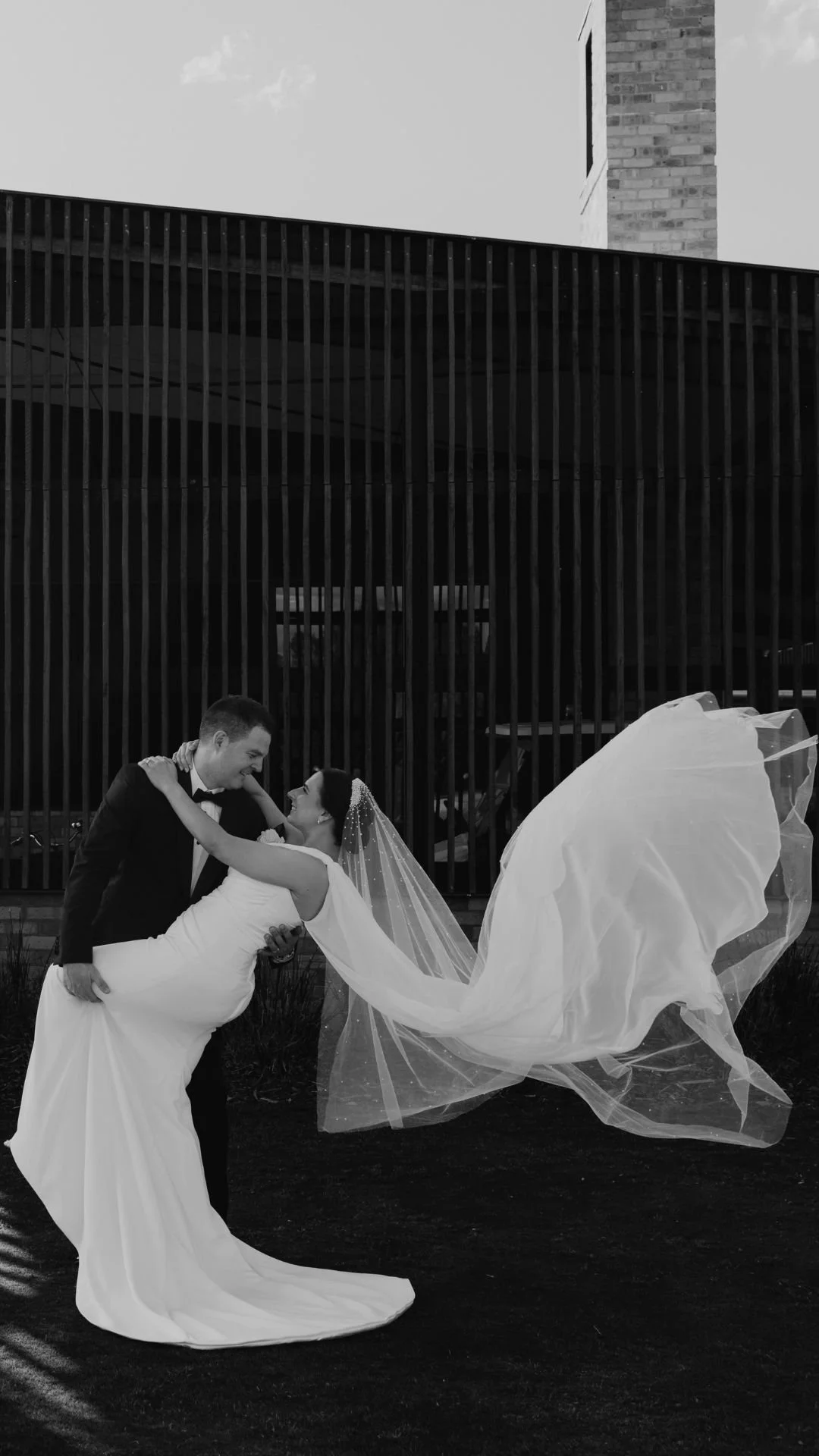A bride and groom sharing a dance outside, with the groom dipping the bride. The bride is wearing a wedding dress and veil, and the groom is dressed in a tuxedo, against a background of a modern building with vertical slats.