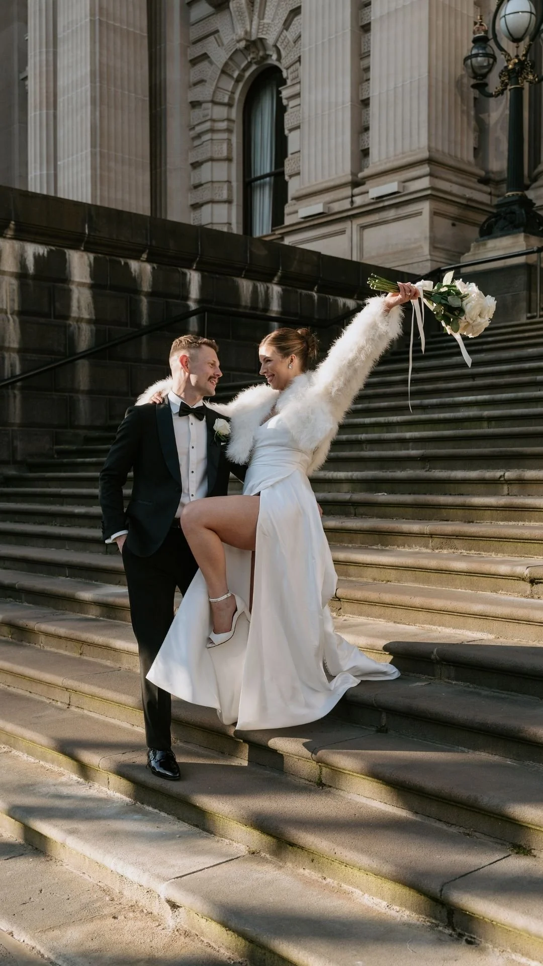 A bride in a white wedding dress and a groom in a black tuxedo pose on stone steps outside a historic building. The bride holds a bouquet of white flowers and has one leg raised, showing her white high-heeled shoe. Both are smiling and looking at eac