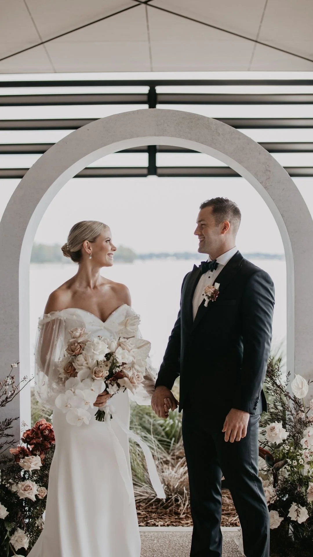 A bride and groom holding hands during their wedding ceremony, standing in front of a scenic view by the water, framed by an archway decorated with flowers.