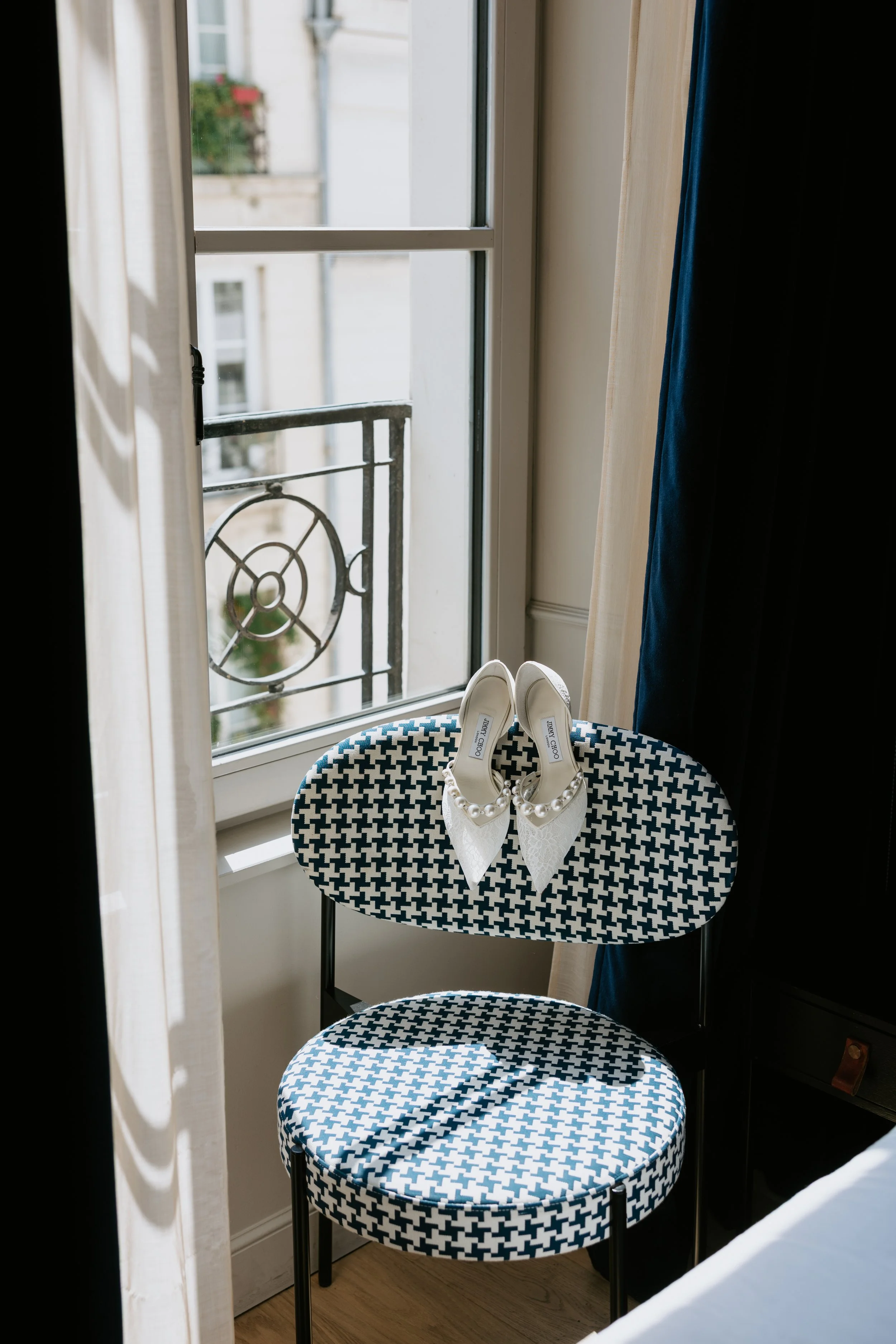 White high-heeled shoes with pearl embellishments on a black and white patterned round table, in front of a window with a black metal balcony railing and a view of neighboring buildings.