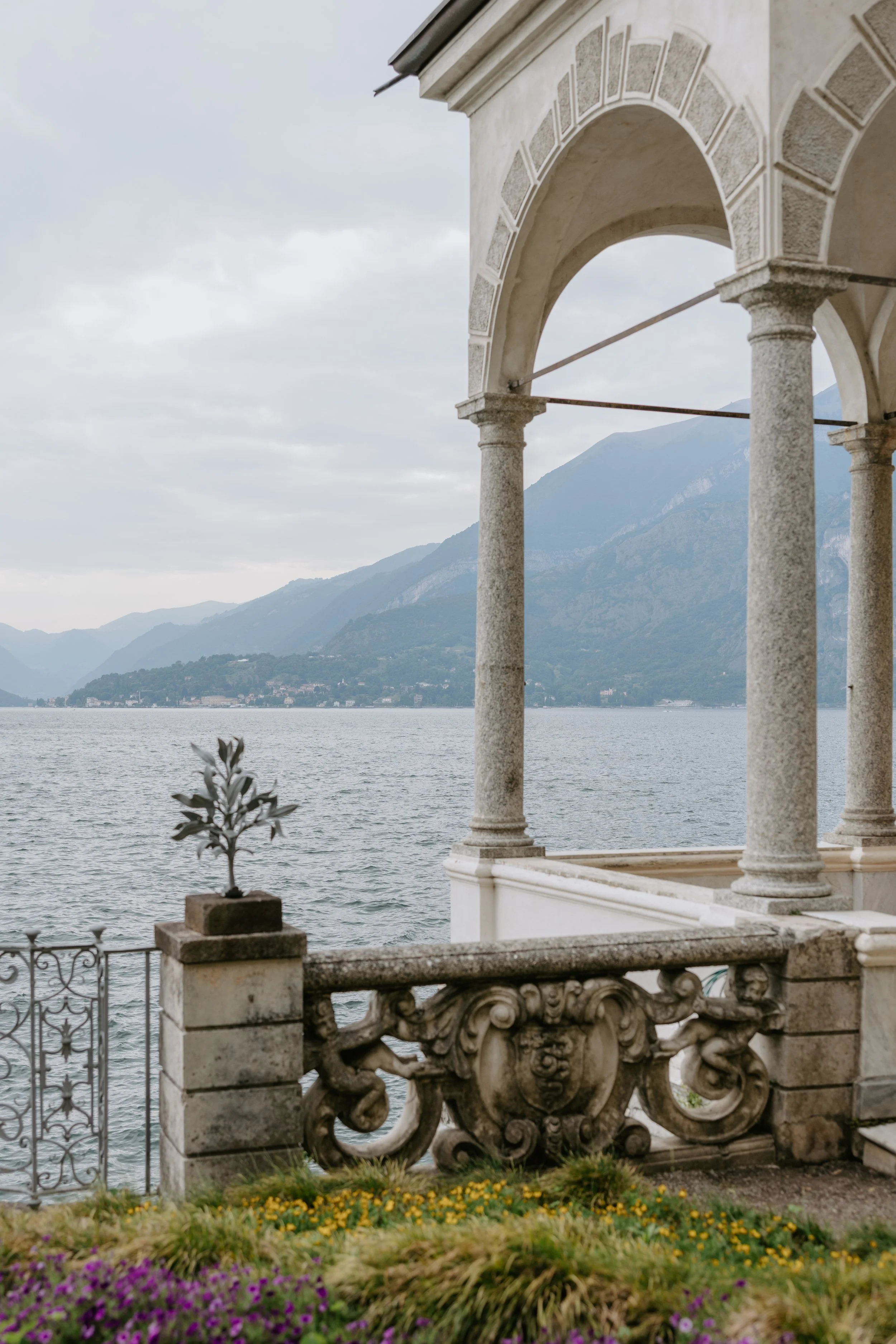 View of a lakeside balcony with ornate stone and metal railing, potted plants, and a view of water and mountains in the distance under a cloudy sky.