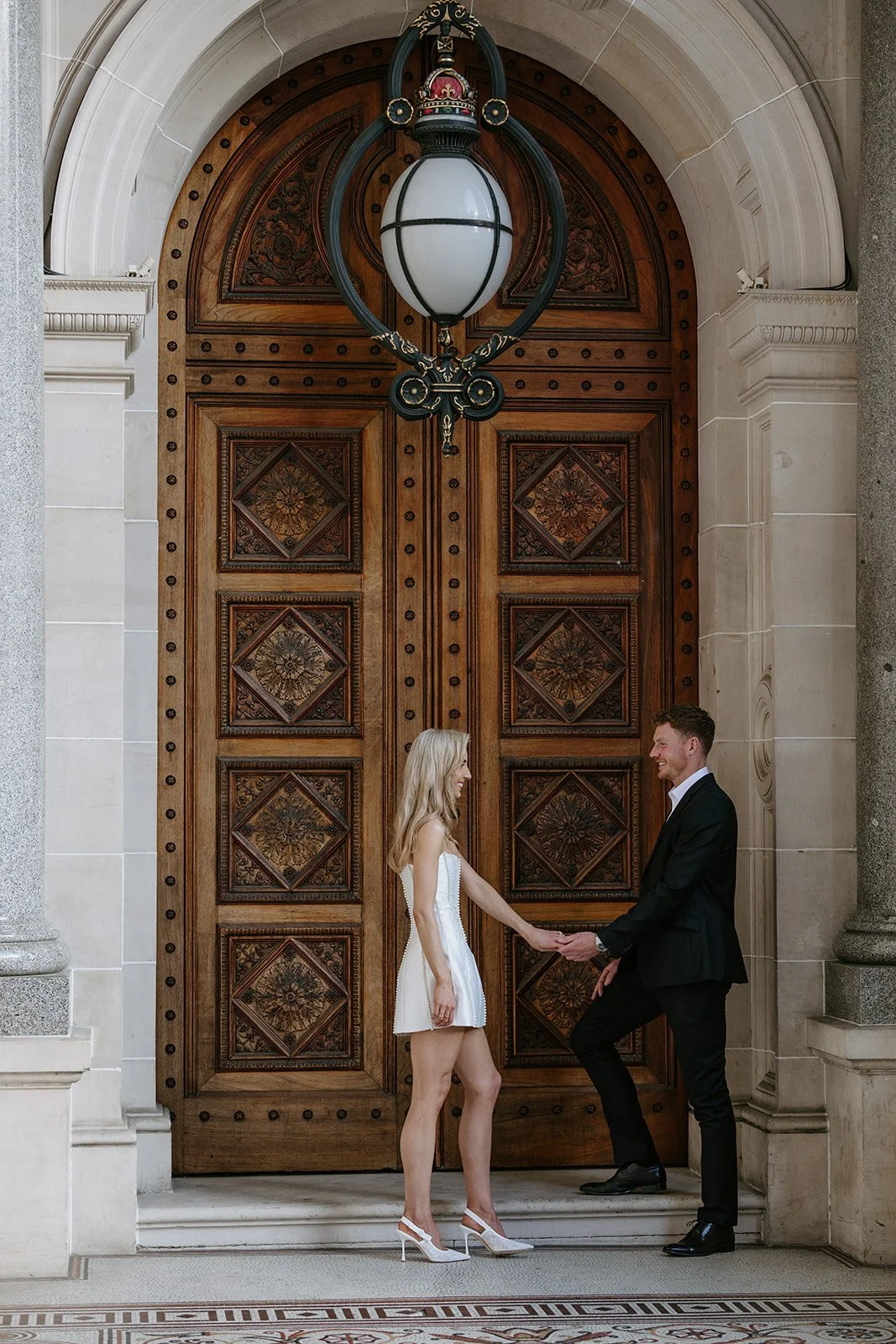 A couple holding hands in front of a large, ornate wooden door with intricate carvings, above a stone steps and columns, hanging from the ceiling is an elegant lantern light fixture.
