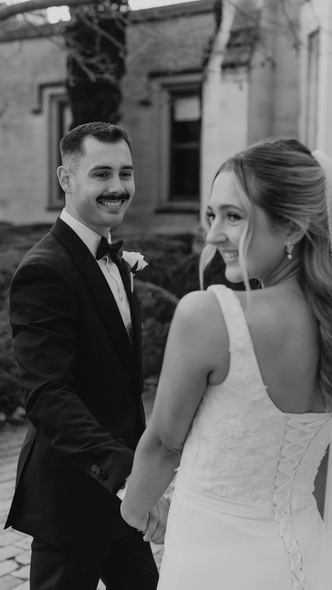 A black and white photograph of a bride and groom holding hands, smiling at each other outdoors, with a building in the background.