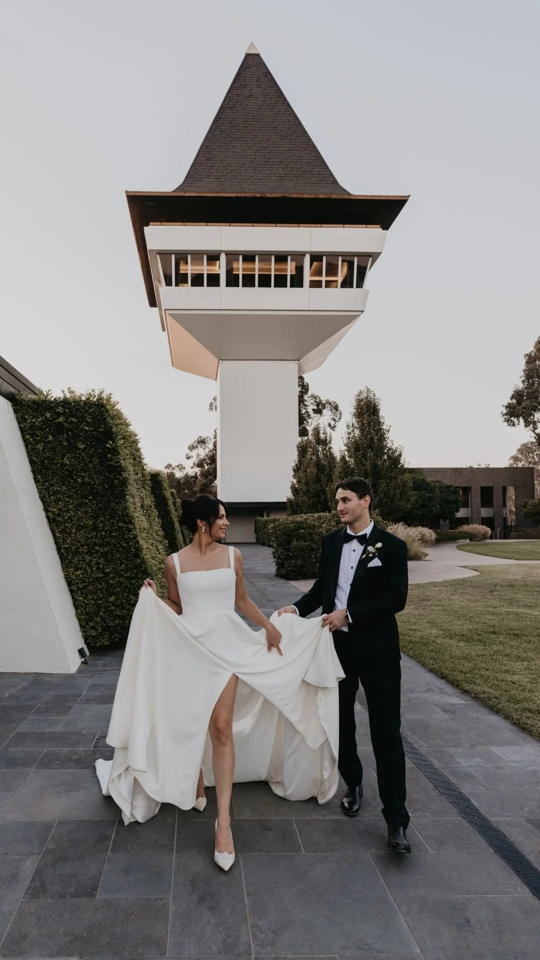 A bride in a white wedding dress and a groom in a tuxedo walking outdoors, holding the bride's dress, with a modern architectural building in the background.