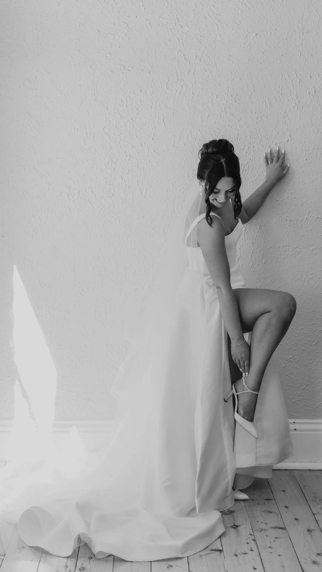 A woman in a wedding dress and high heels, smiling and posing against a textured white wall, with her right hand on wall and left hand on bent knee, facing slightly away from camera.