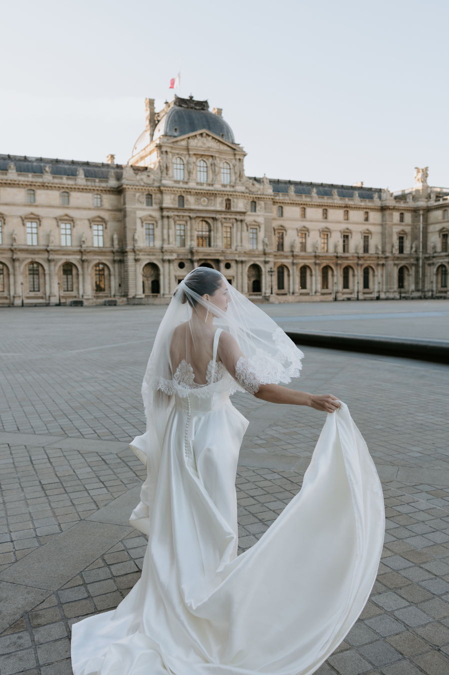 A bride in a white wedding dress and veil holding the train with a historic European building in the background.