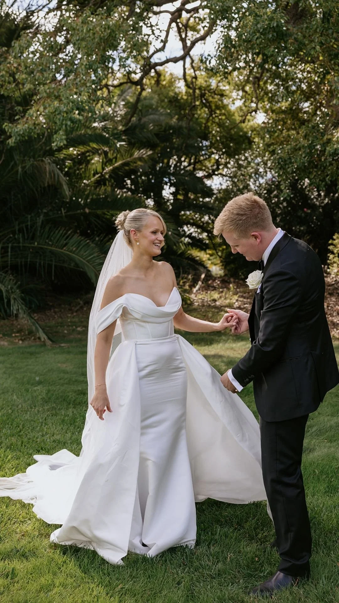 Bride and groom holding hands outdoors on green grass with trees in the background, smiling and dressed in wedding attire.