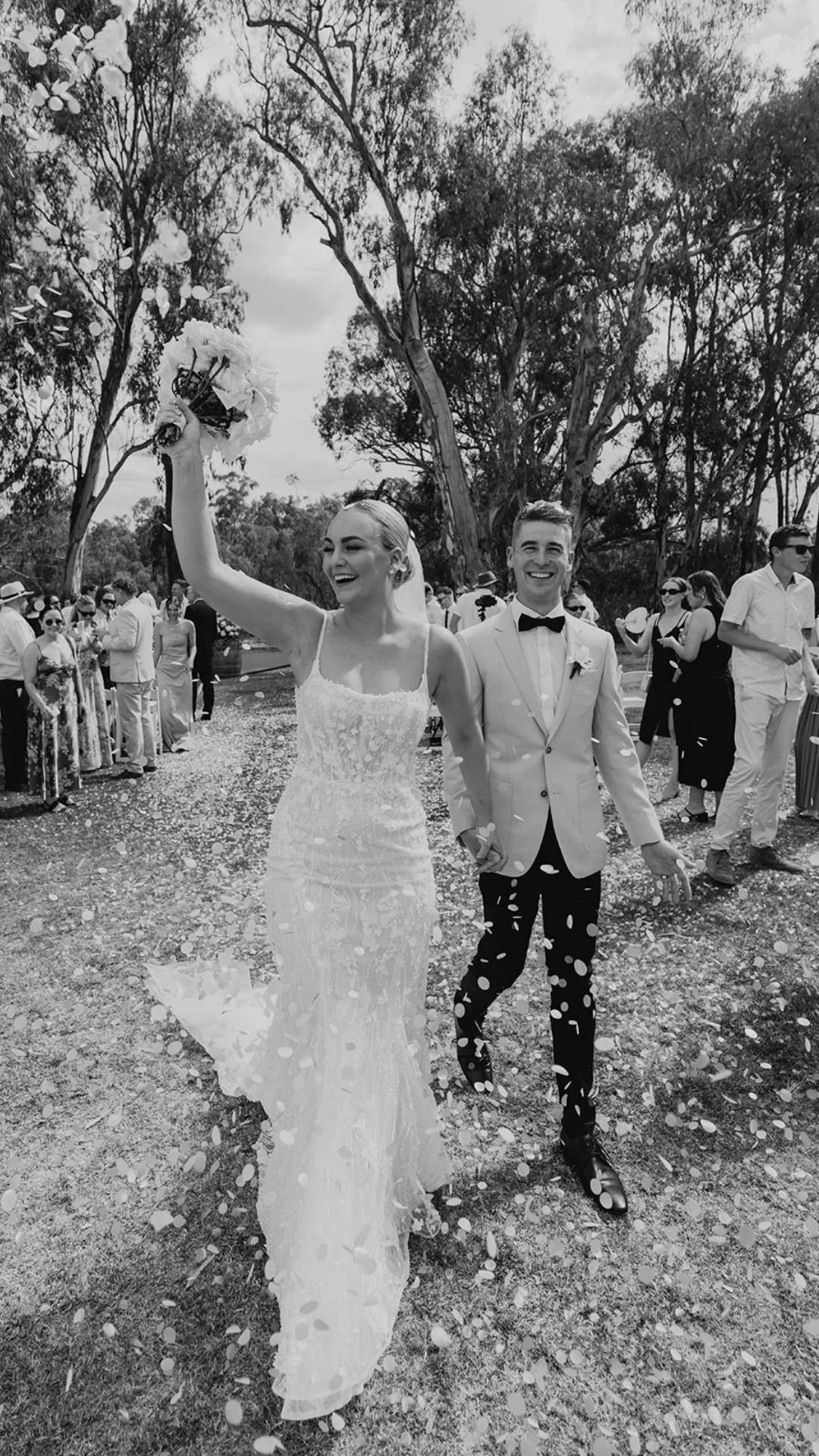 A bride and groom walking outdoors after their wedding, surrounded by guests and throwing confetti, with trees in the background.