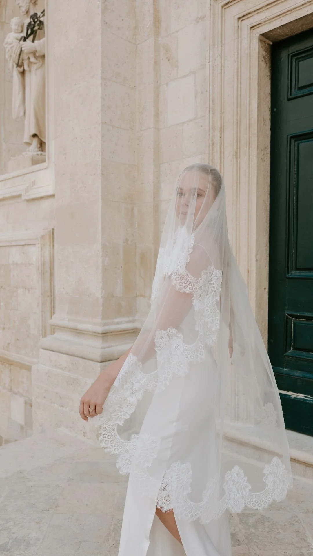A woman wearing a white lace wedding dress and veil standing outside near a beige stone wall and green door.