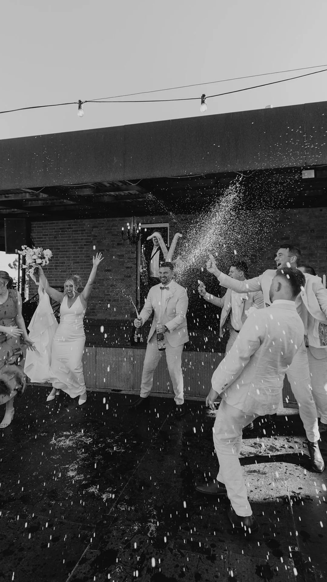People celebrating at a wedding reception, throwing rice, dressed in formal attire, with a brick wall background and string lights overhead.