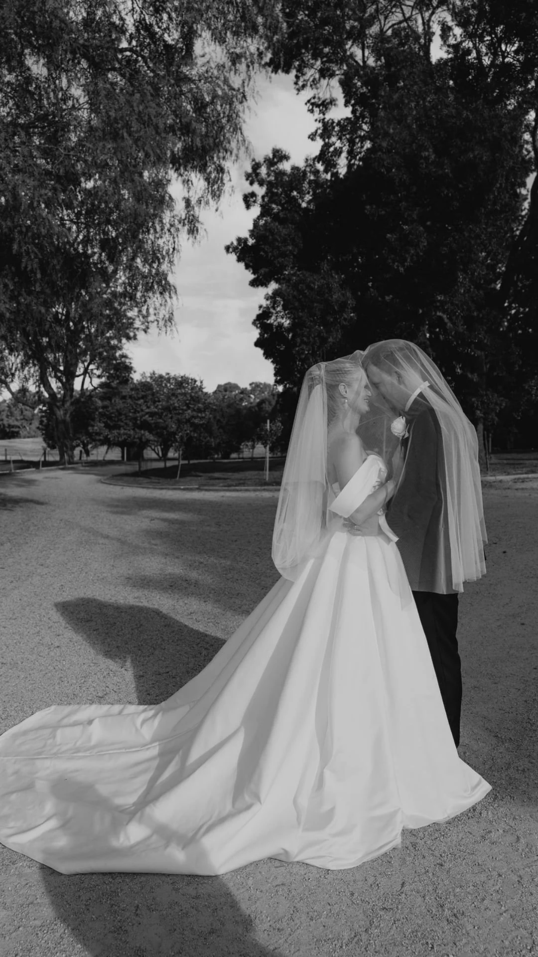 Black and white photo of a bride and groom standing close together outdoors under a veil. The bride wears a wedding gown, and the groom wears a tuxedo with a boutonniere. They are smiling and touching foreheads, surrounded by trees and a cloudy sky.