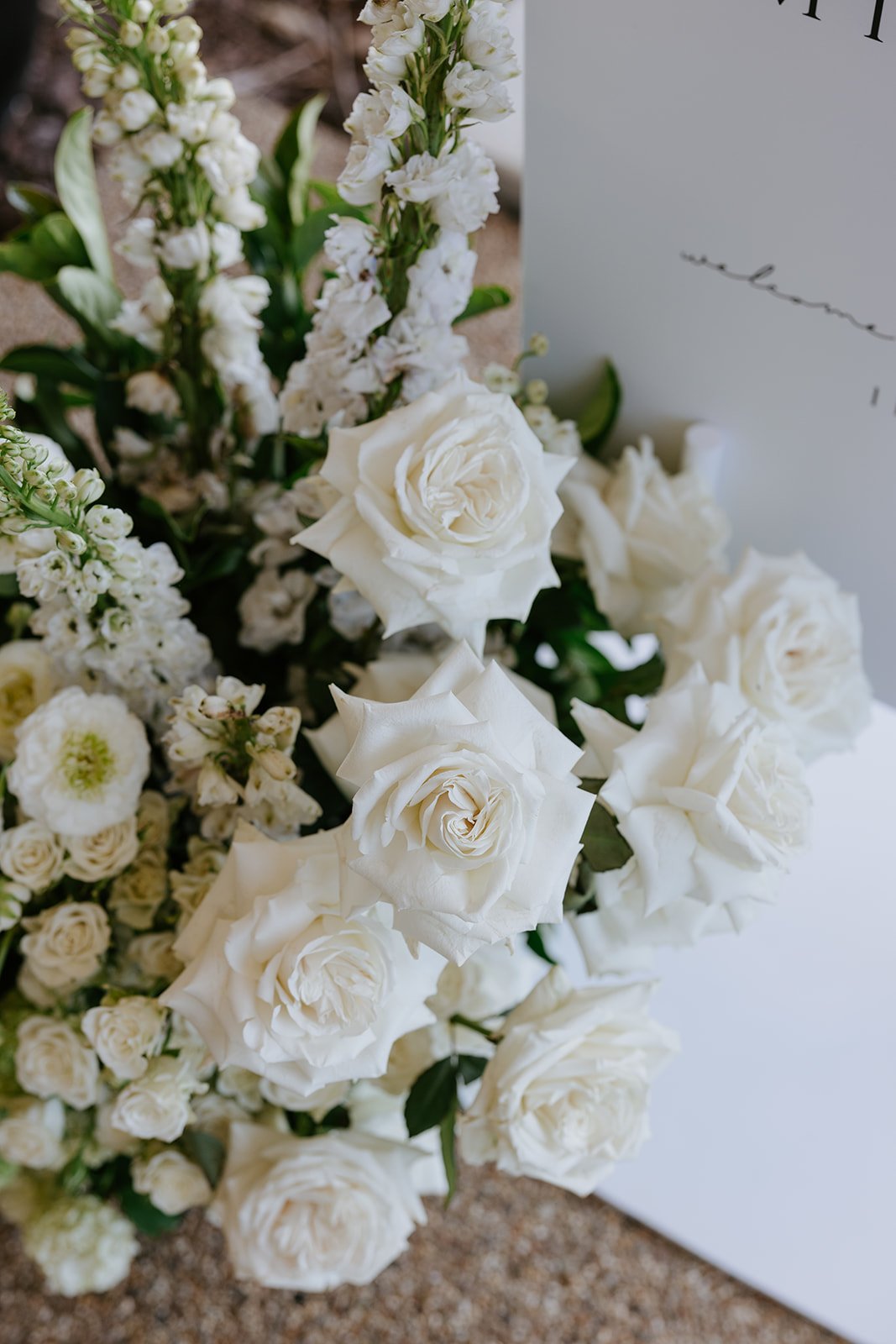 Close-up of a white floral bouquet with roses and other white flowers, with a part of a white card or sign in the background.