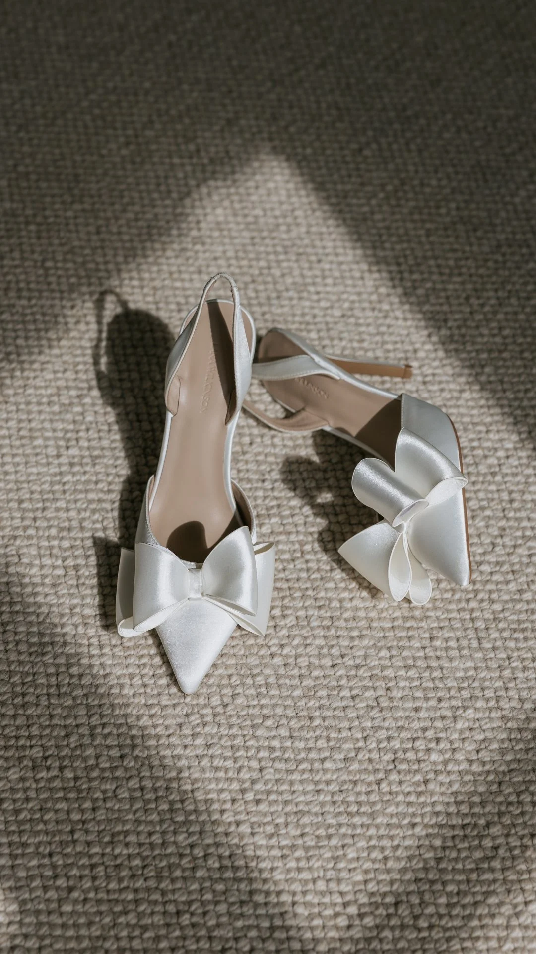 White high-heeled shoes with large bows on the pointed toes, placed on a beige textured carpet, with sunlight casting a shadow.