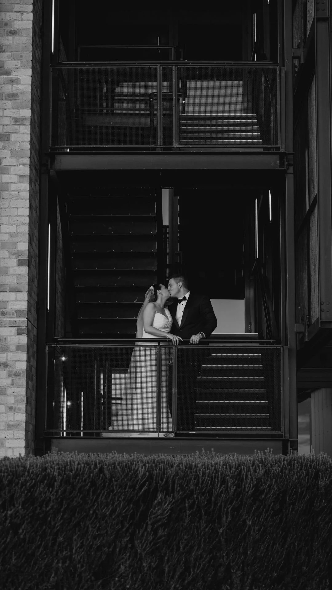Black and white photo of a bride and groom sharing a kiss on a building's fire escape balcony.