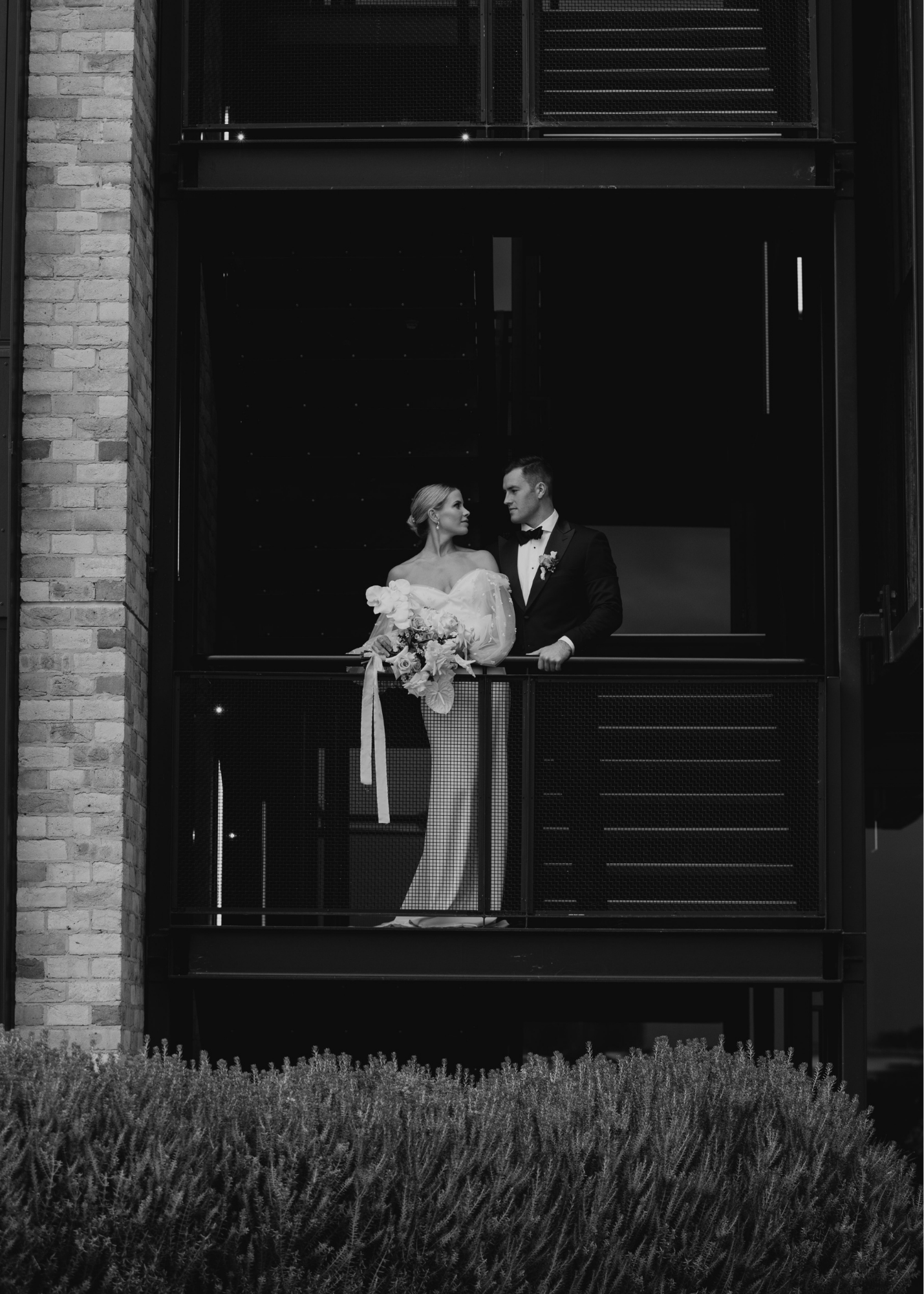 Black and white photo of a bride and groom standing on a balcony, looking at each other, with the bride holding a bouquet. The balcony is part of a modern building with industrial-style metal and brickwork.