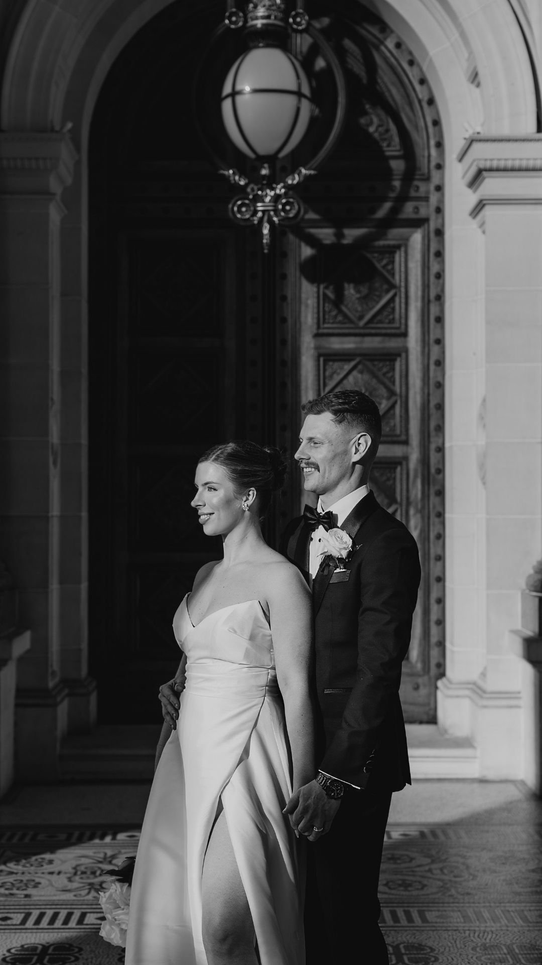 Black and white photo of a bride and groom standing together in a grand indoor setting, smiling and dressed in wedding attire.
