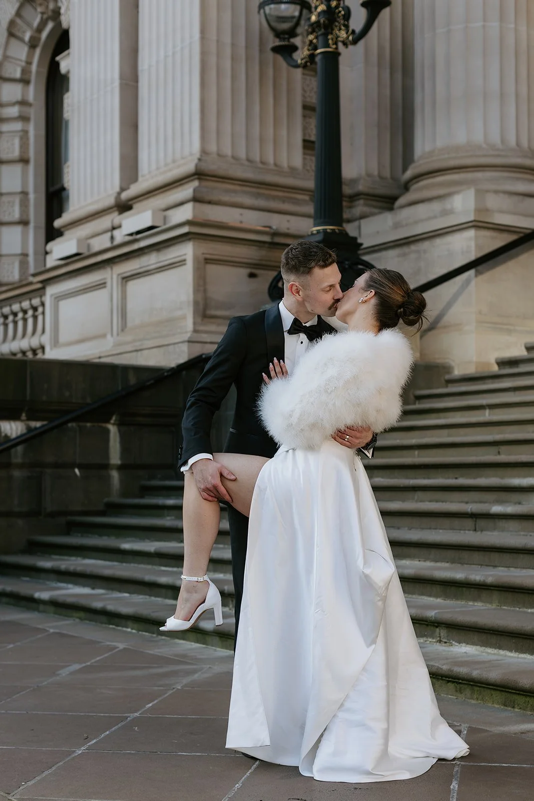 A bride and groom share a kiss on the stairs outside a historic building. The groom, in a tuxedo, holds the bride, who is wearing a white wedding dress and high heels, while she has a white fur stole around her shoulders.