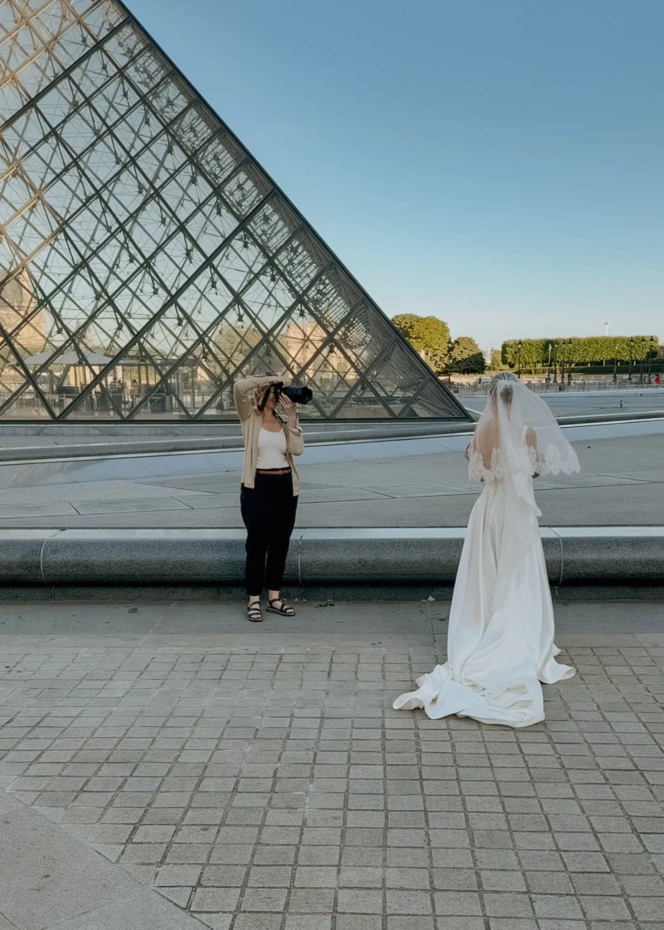 A woman in a beige jacket and black pants taking a photograph of a bride in a white wedding dress with a long veil, standing outdoors near a glass pyramid structure and trees.