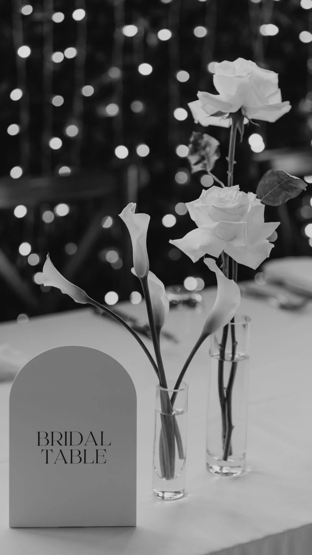 Three vases with white flowers on a table, with a 'BRIDAL TABLE' sign and blurred lights in the background.