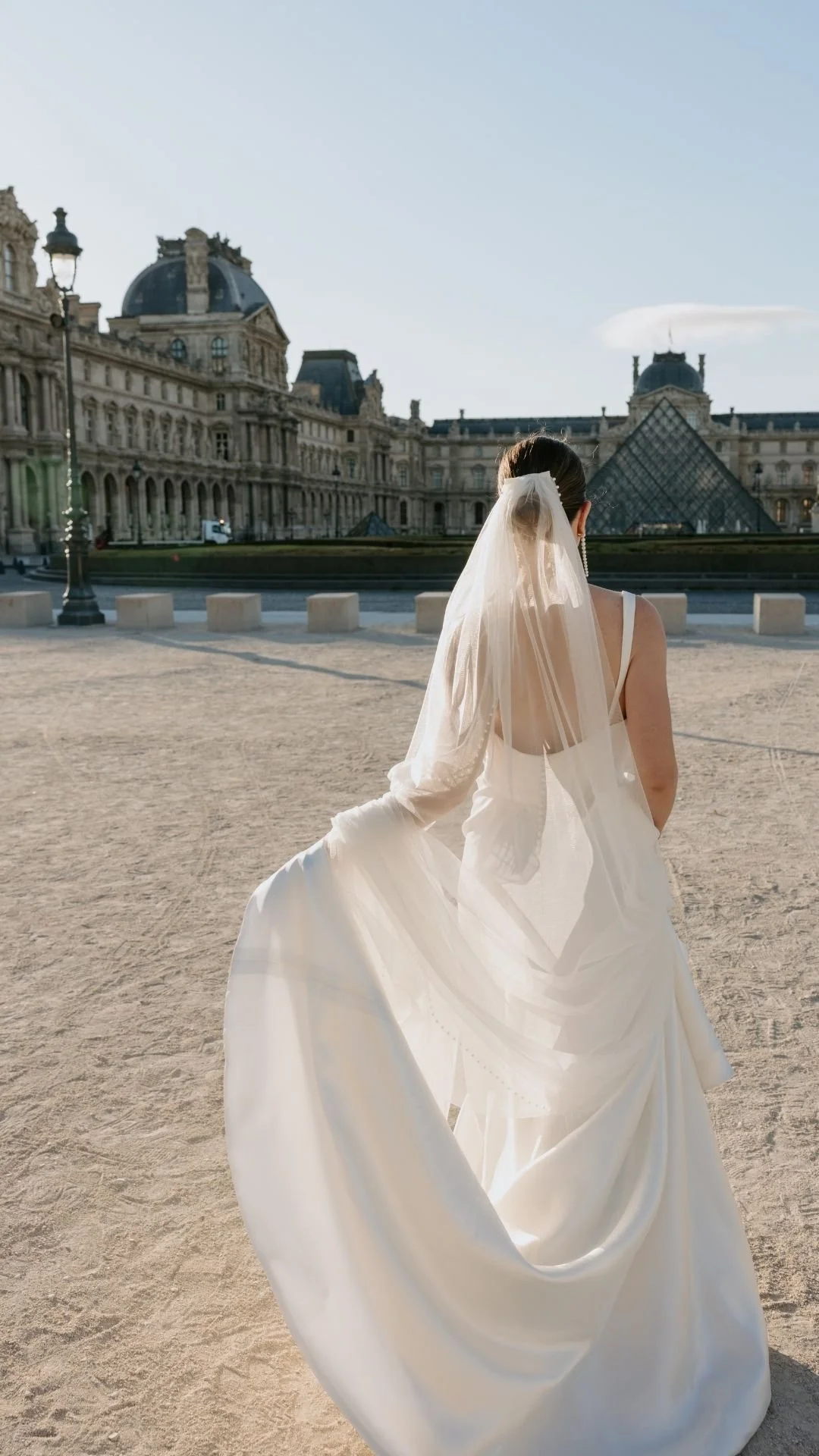 Woman in a white wedding dress with a long veil, sitting on a sandy surface in front of the Louvre Museum in Paris, France.