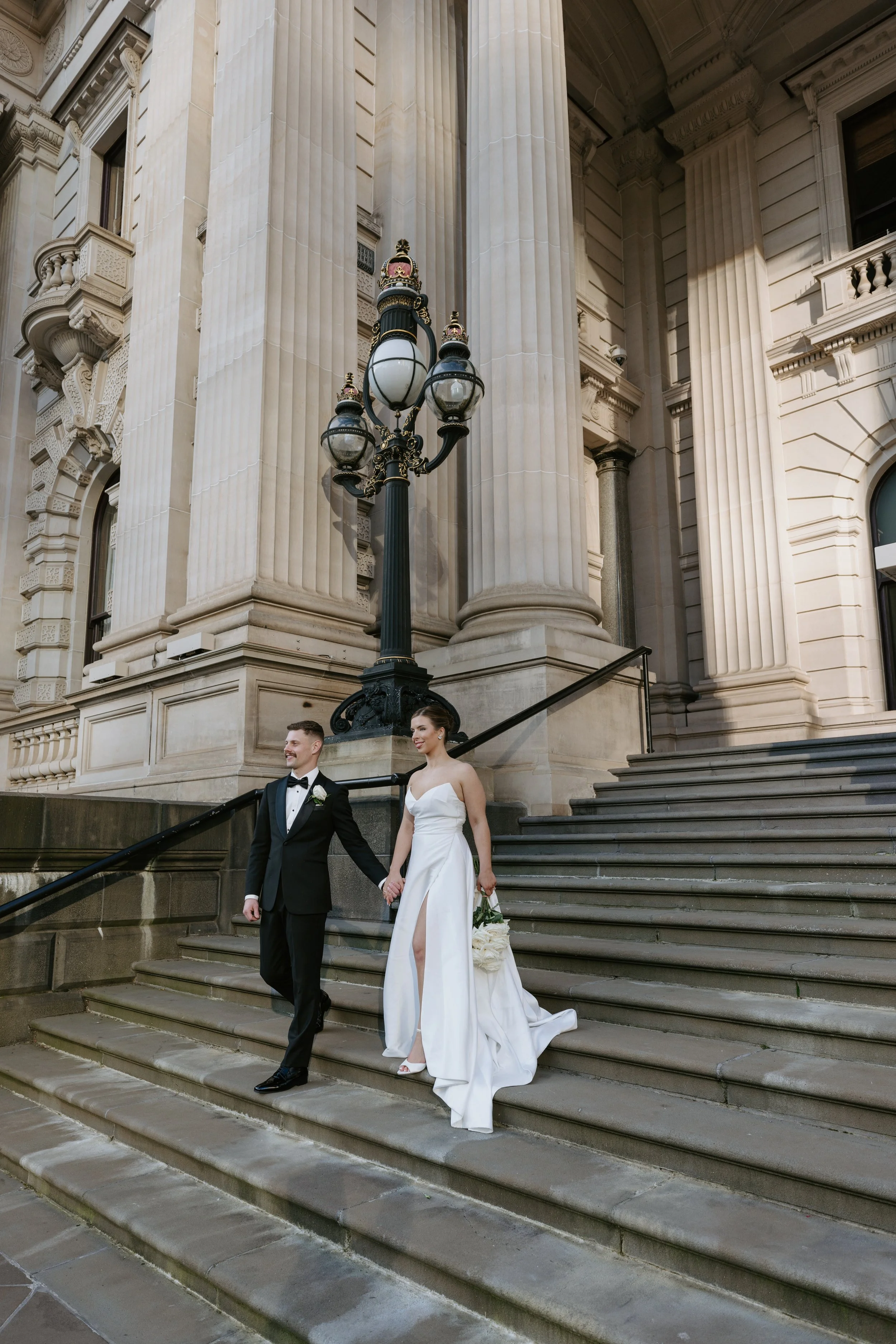 Newlywed couple walking down the steps of Parliament House in Melbourne, captured in an elegant editorial wedding photography by love by shae.