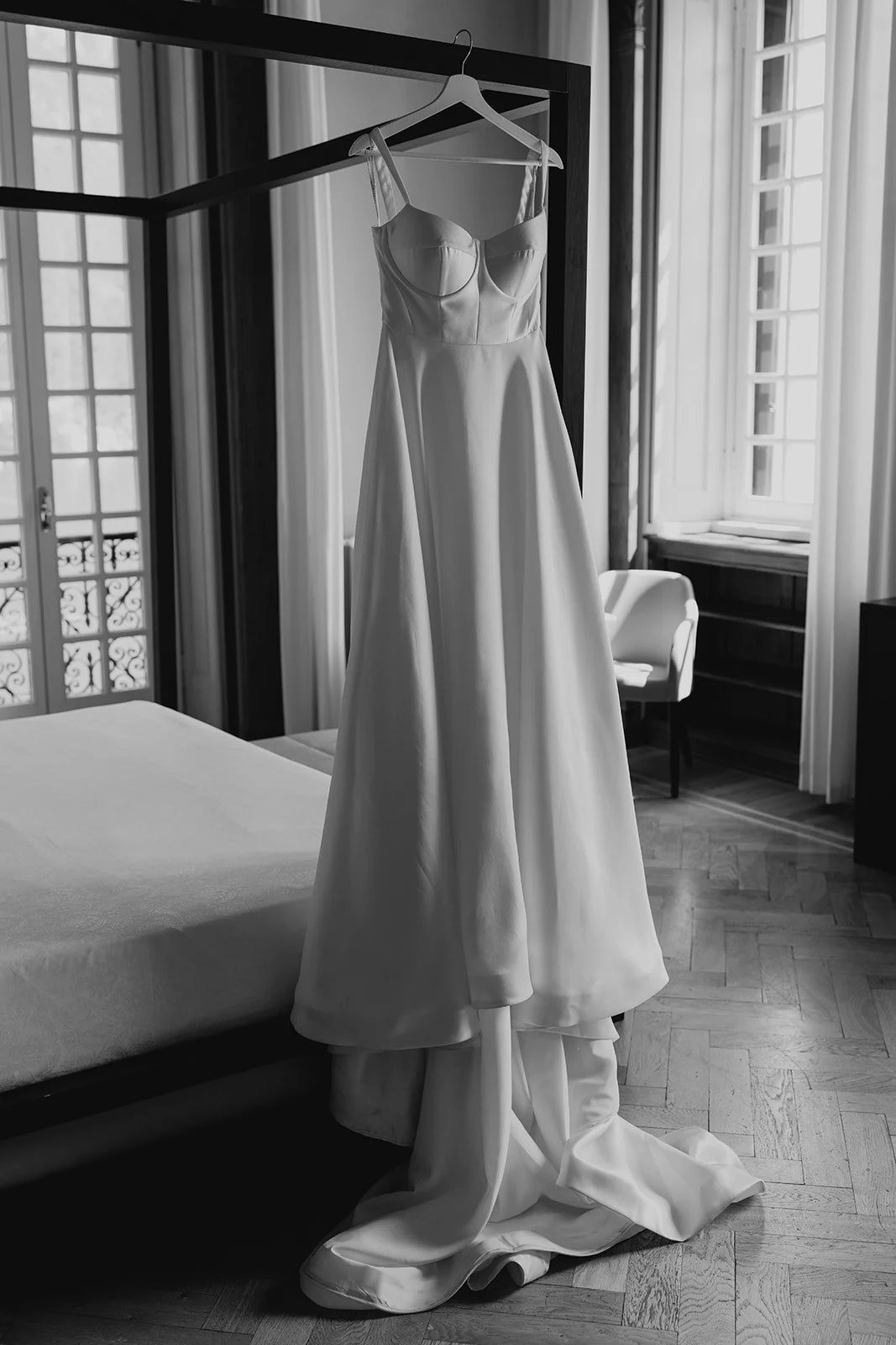 A white wedding dress hanging on a hanger on a four-poster bed in a bright, elegantly furnished room.