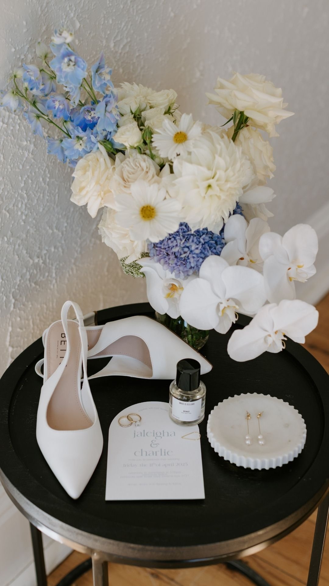 A black circular table with white high-heeled shoes, a perfume bottle, wedding rings, a floral invitation, a white jewelry dish with earrings, and a large bouquet of white, blue, and purple flowers against a textured white wall.