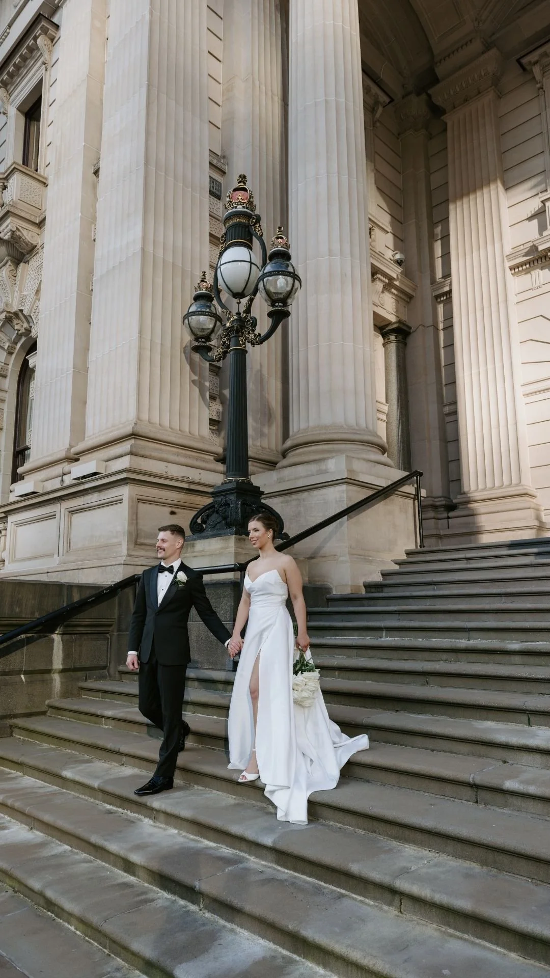 A newlywed couple walking down stone steps outside a historic building with large columns and ornate architectural details. The groom is in a black tuxedo with a bow tie, and the bride is in a white wedding gown holding a bouquet of white flowers.