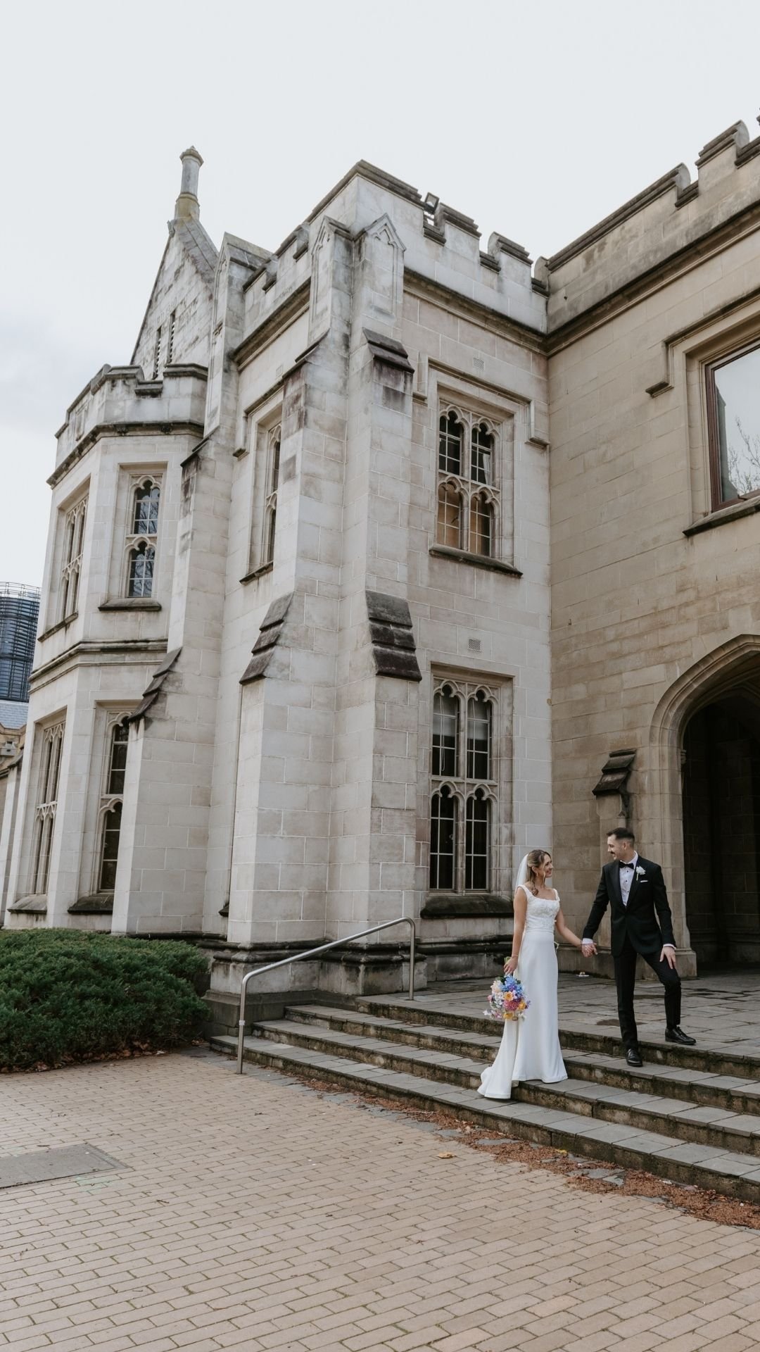 A bride and groom holding hands on the steps of a historic stone building with Gothic-style windows. The bride is in a white wedding dress holding a colorful bouquet, and the groom is in a black tuxedo.