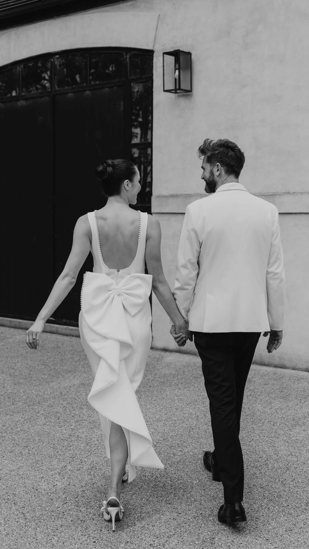A couple walking hand in hand dressed in formal attire with the woman in a white gown and the man in a white jacket holding hands and smiling in European Wedding Venue with Melbourne wedding photographer love by shae.