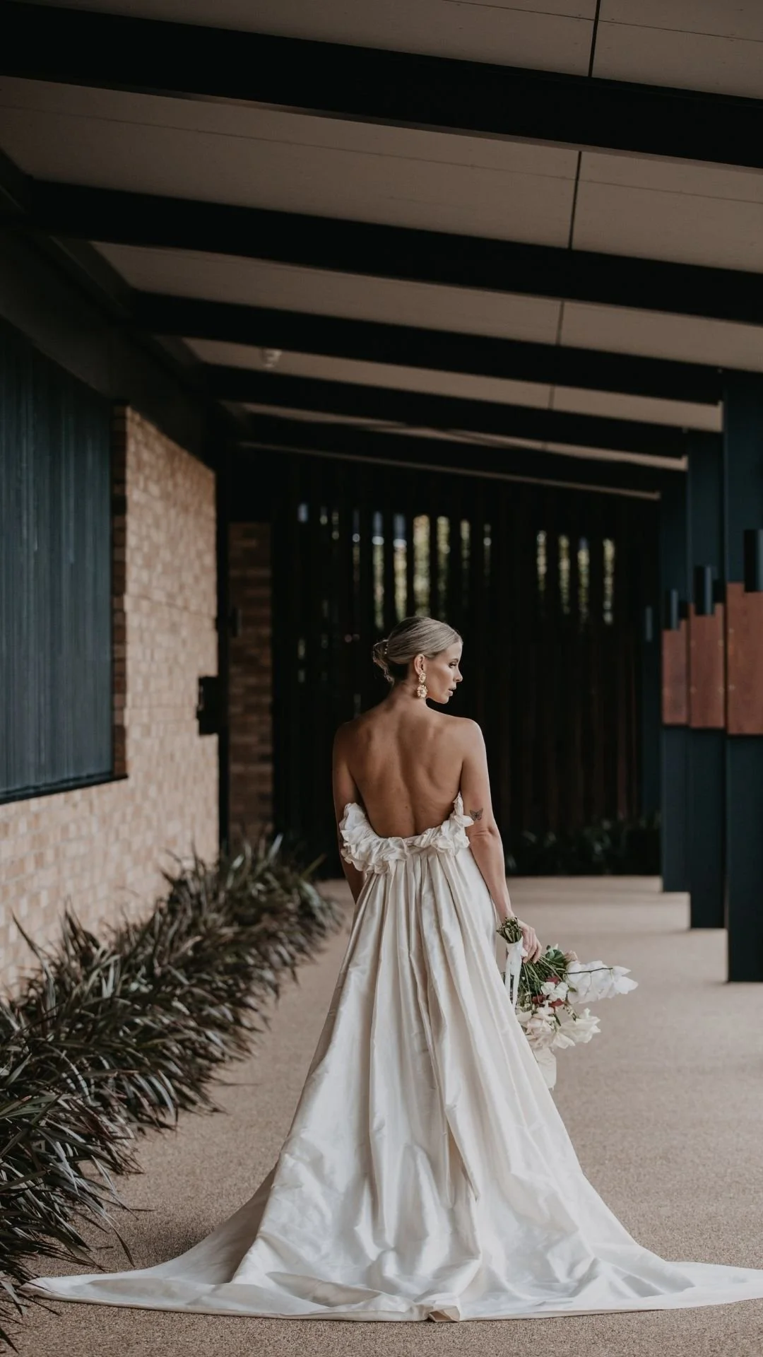 A woman in a backless wedding dress holding a bouquet standing outdoors near a modern building with brick and metal accents.