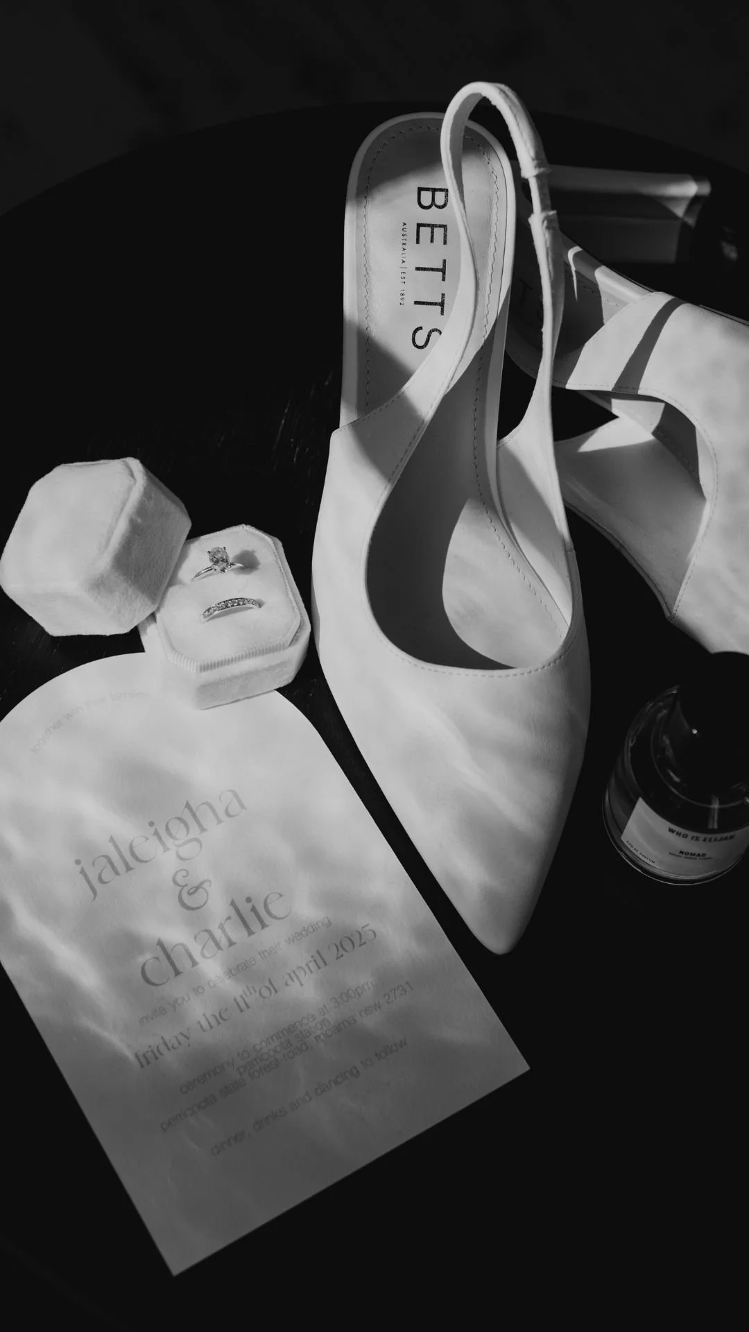 Wedding invitation, white high heel shoes, diamond ring in a box, perfume bottle, and a pink ring box on a dark surface.