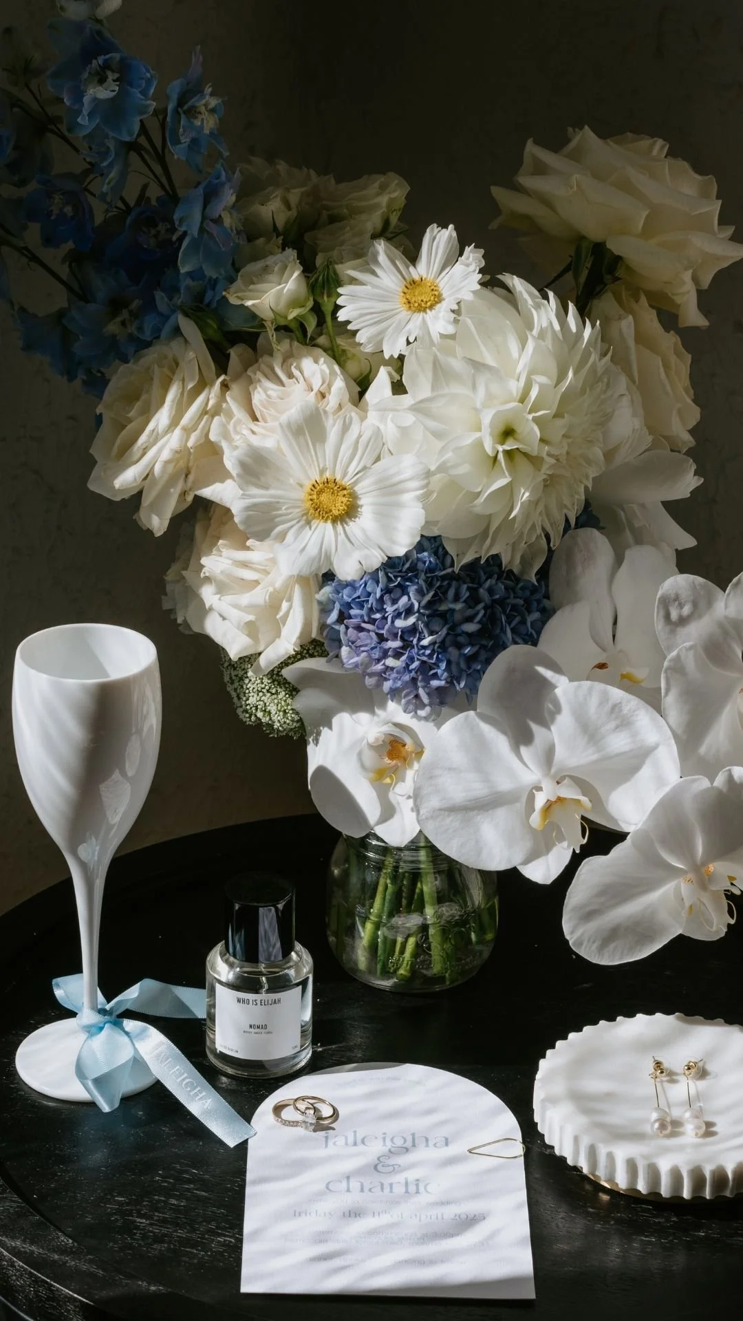 A floral wedding arrangement with white, blue, and cream-colored flowers, a white ceramic vase, a perfume bottle, a white ribboned champagne flute, wedding rings, and a printed wedding invitation on a black surface.